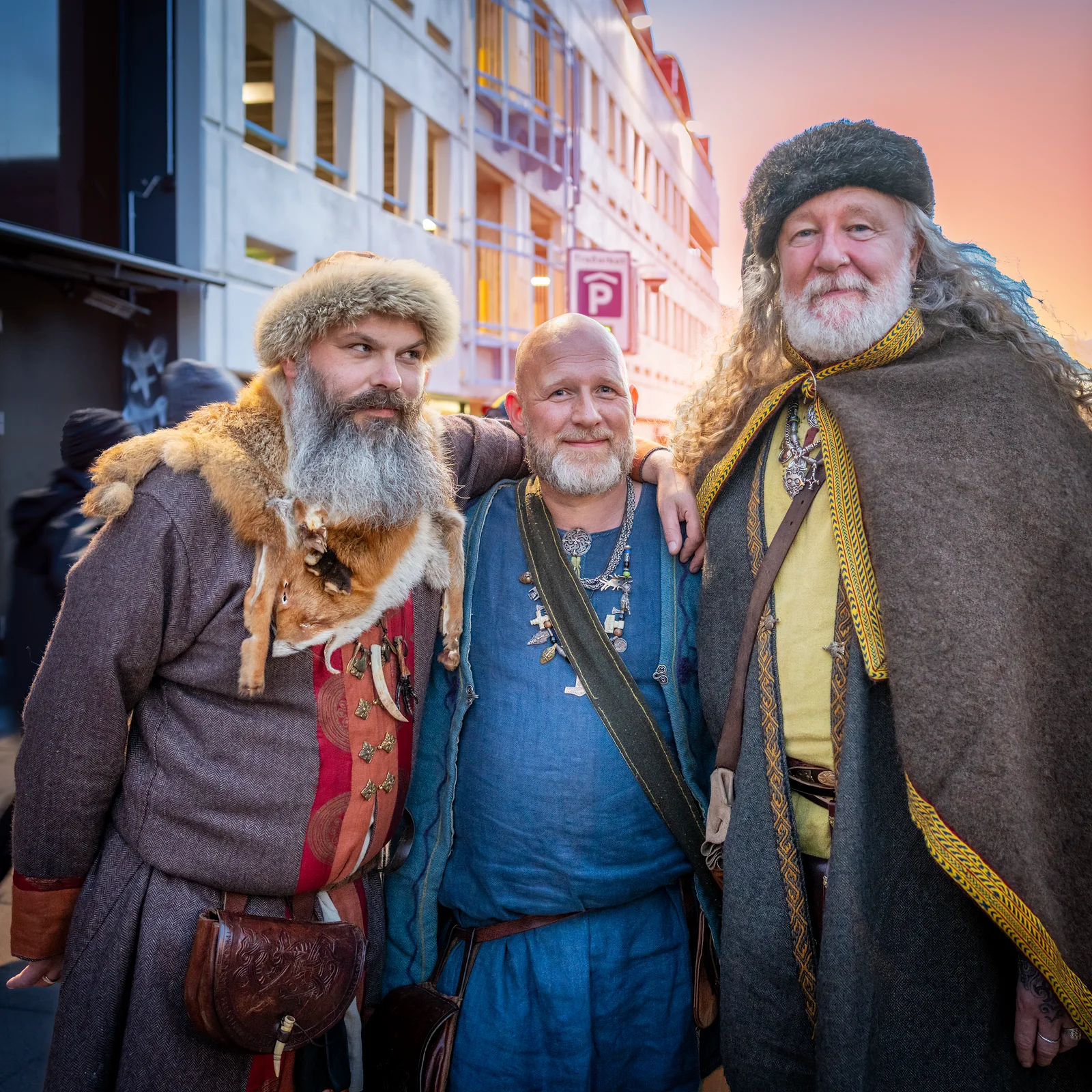 Here are three alt text variations tailored to fit your layout requirements, strictly adhering to the character limit constraints and accessibility guidelines. Option 1: Clean & Factual (Recommended) Three men in detailed, traditional Viking-age attire stand arm-in-arm on a city street during Reykjavík Culture Night. They wear historical clothing, including tunics, fur trims, heavy cloaks, and historical jewelry, smiling under a sunset sky.