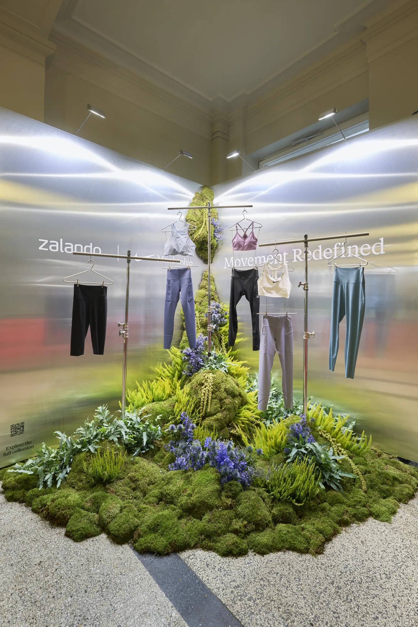 Retail display of Zalando × Nike sportswear hanging on clothing rails, presented on a hill of moss, ferns, and blue flowers in a polished metallic setting.