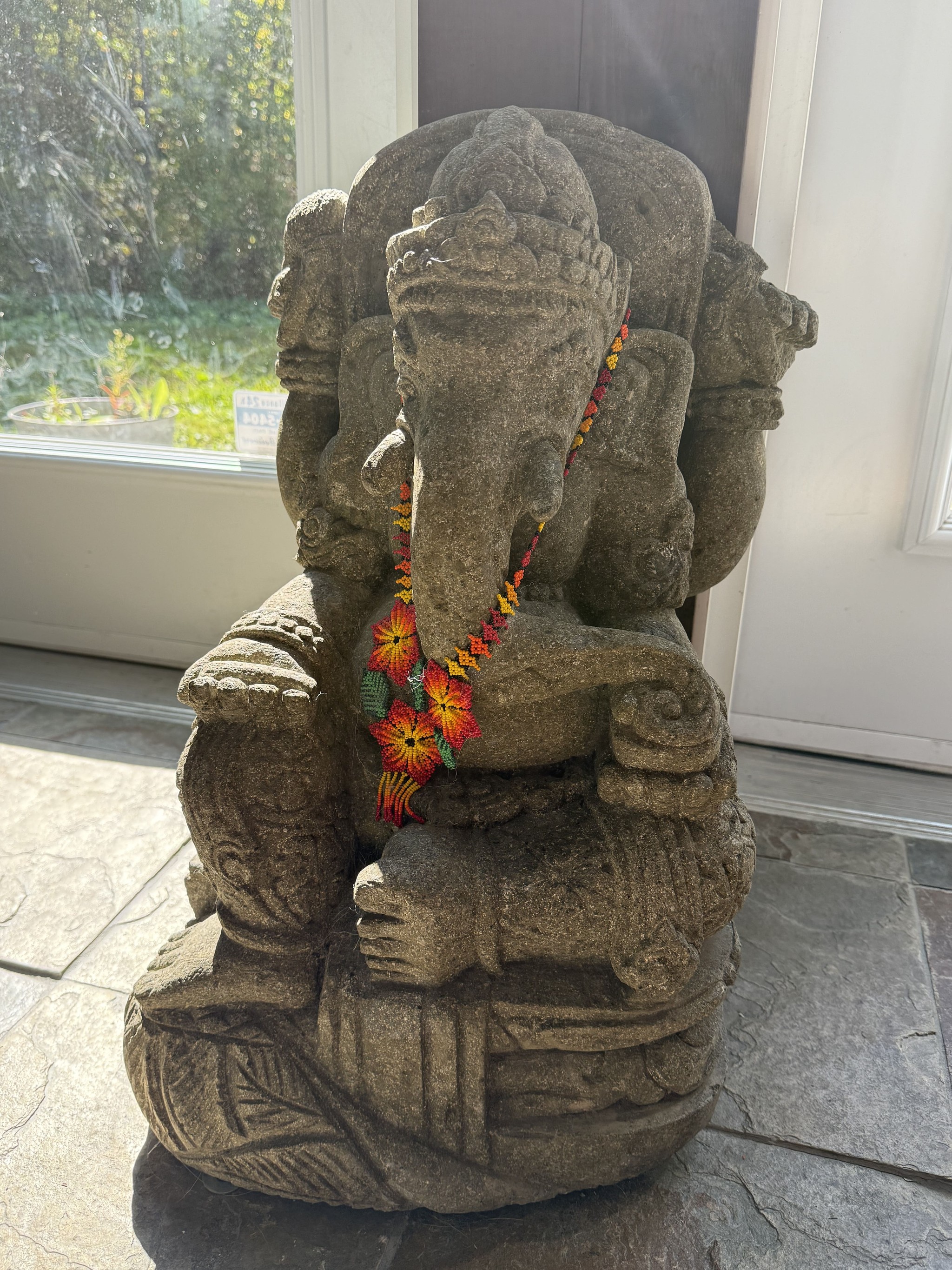 Sculpture en pierre de la divinité hindoue ornée d’un collier de fleurs colorées près d’une baie vitrée ensoleillée.