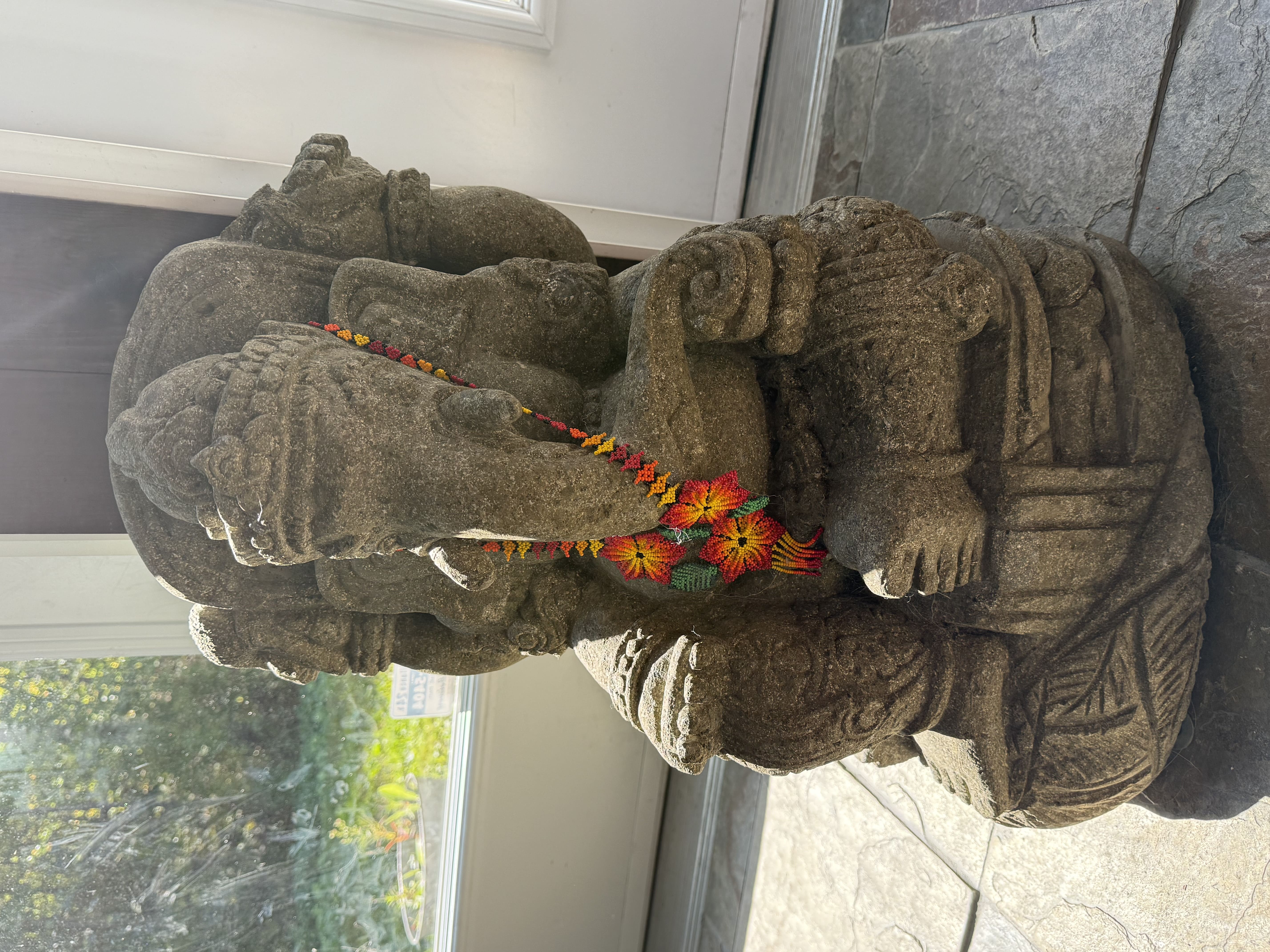 Sculpture en pierre de la divinité hindoue ornée d’un collier de fleurs colorées près d’une baie vitrée ensoleillée.