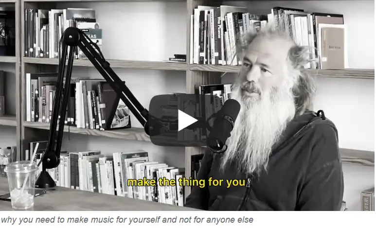 Man with a beard speaking into a microphone, discussing creativity and personal expression, surrounded by bookshelves, emphasizing the importance of making music for oneself.