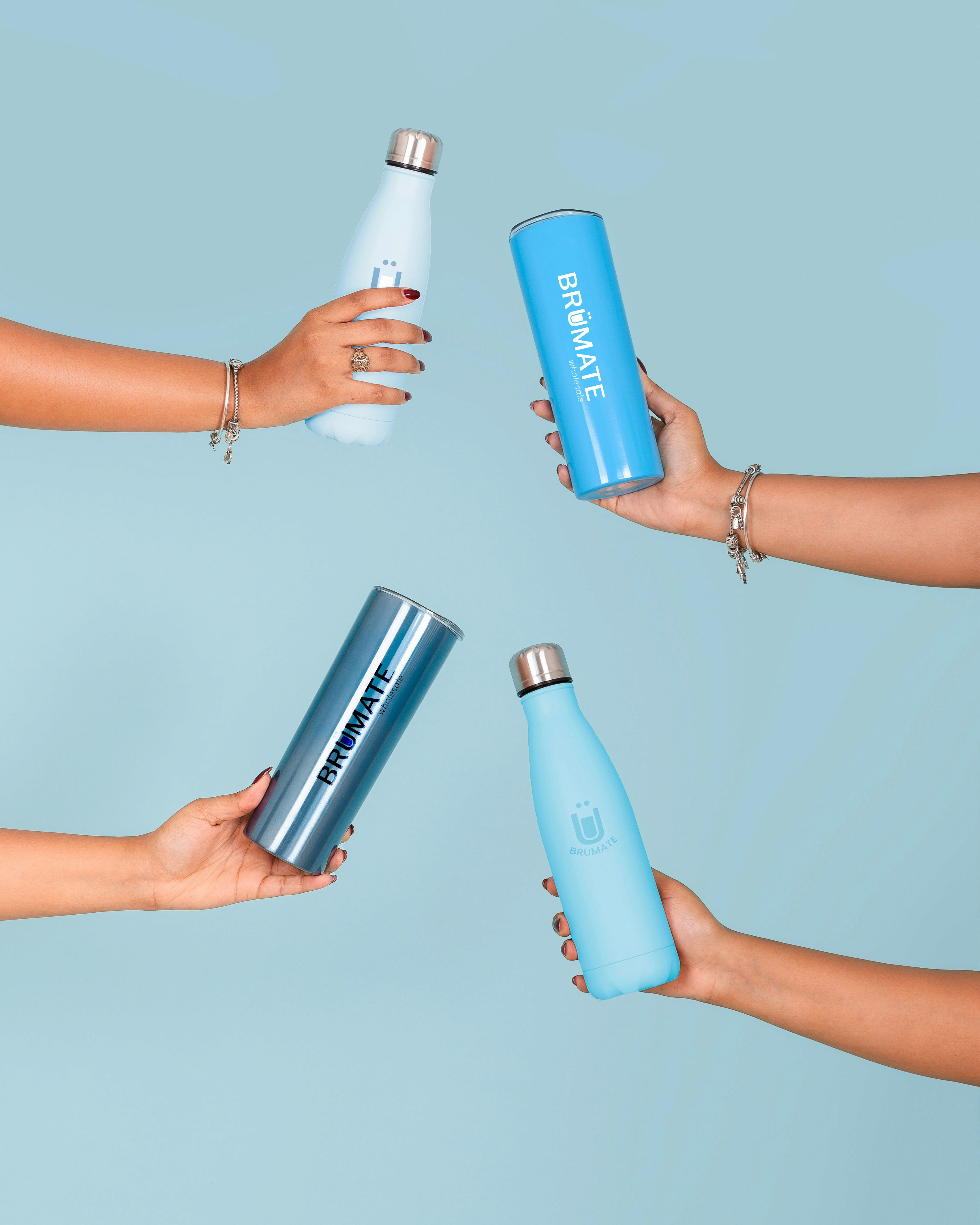 Four hands holding pastel blue insulated bottles and tumblers from BrüMate arranged against a light blue background, creating a clean, minimal product display.