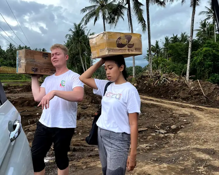 Two volunteers carry boxes of supplies along a rural Bali road to deliver food and training materials