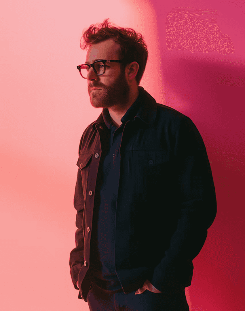A bearded man wearing glasses stands confidently against a vibrant pink and red gradient background, dressed in a dark jacket with hands casually in his pockets.