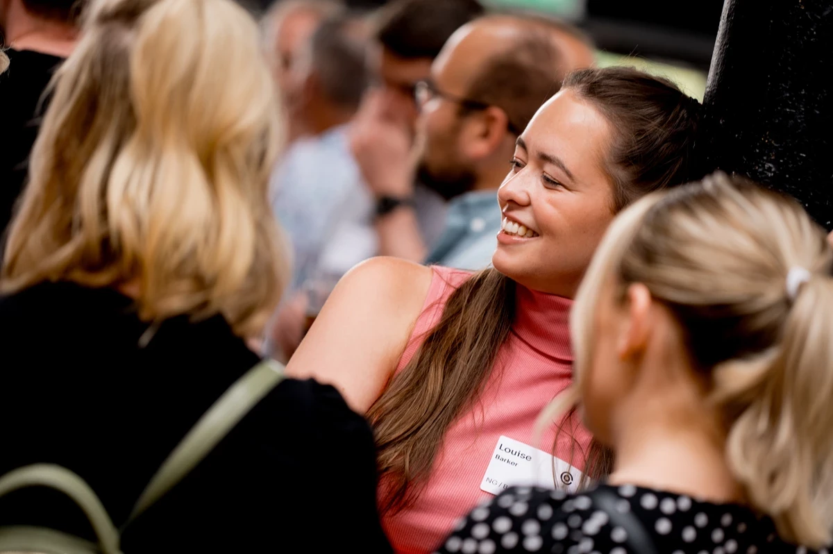 Attendees gather closely in conversation at a networking event or conference. Name badges, casual attire, and a busy background suggest an active professional meetup.