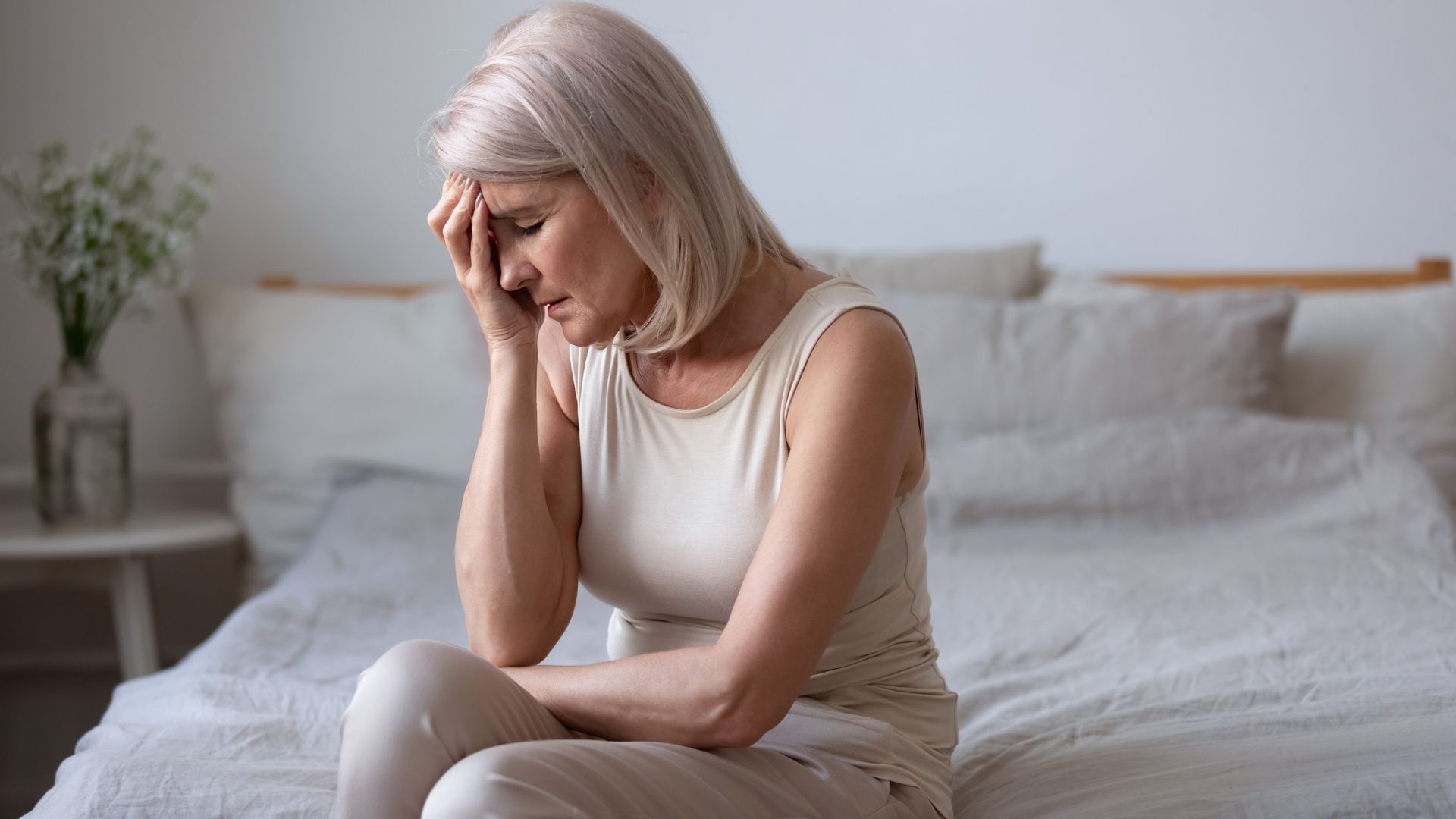 A woman with short blonde hair sits on a bed, resting her forehead on her hand, looking distressed.