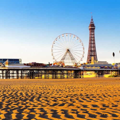 A sandy beach with a Ferris wheel and an Eiffel Tower-like structure on a pier, against a clear blue sky.