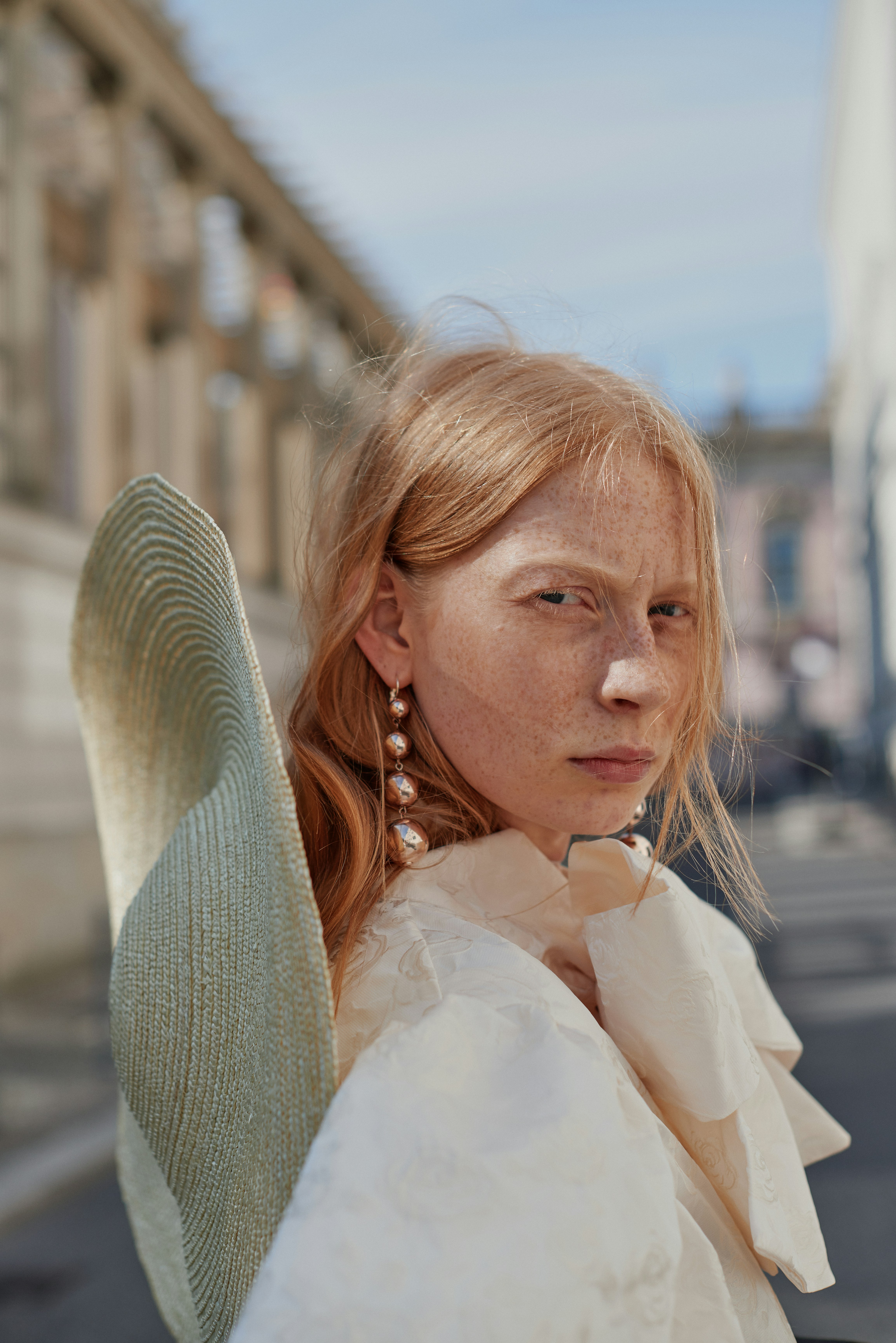 Soft outdoor portrait of a freckled redhead model holding a straw hat, wearing pearl drop earrings and a white ruffled top, architectural background
