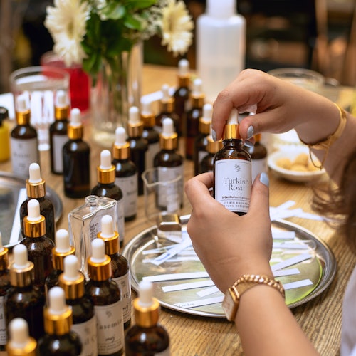 Malapitan na larawan ng mga kamay na may hawak na bote na may label na "Turkish Rose" sa gitna ng iba't ibang sangkap ng pabango sa isang mesa.