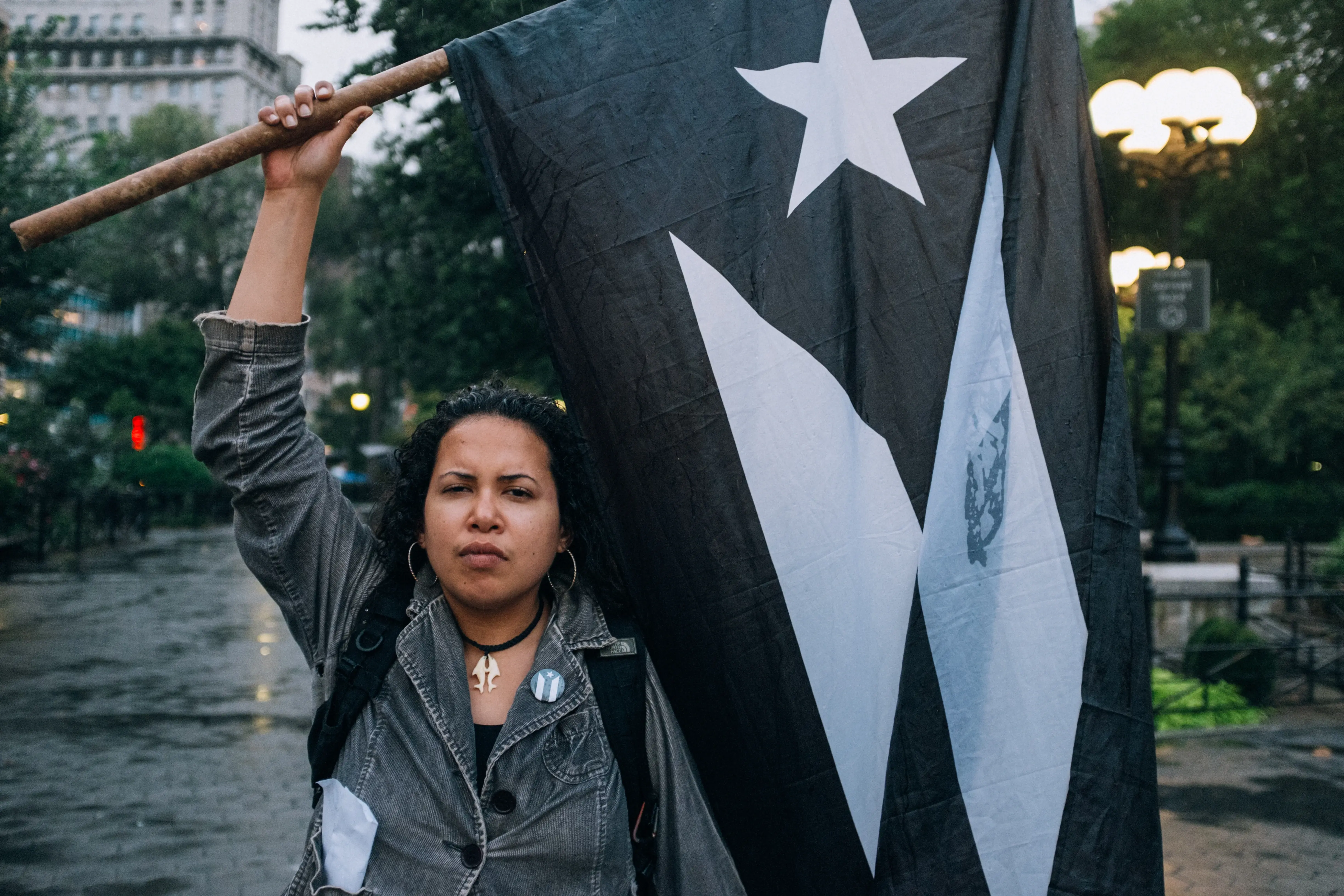 A person stands outdoors holding a black and white flag with a star design, set against a backdrop of trees and buildings in an urban environment, conveying a sense of determination and advocacy.