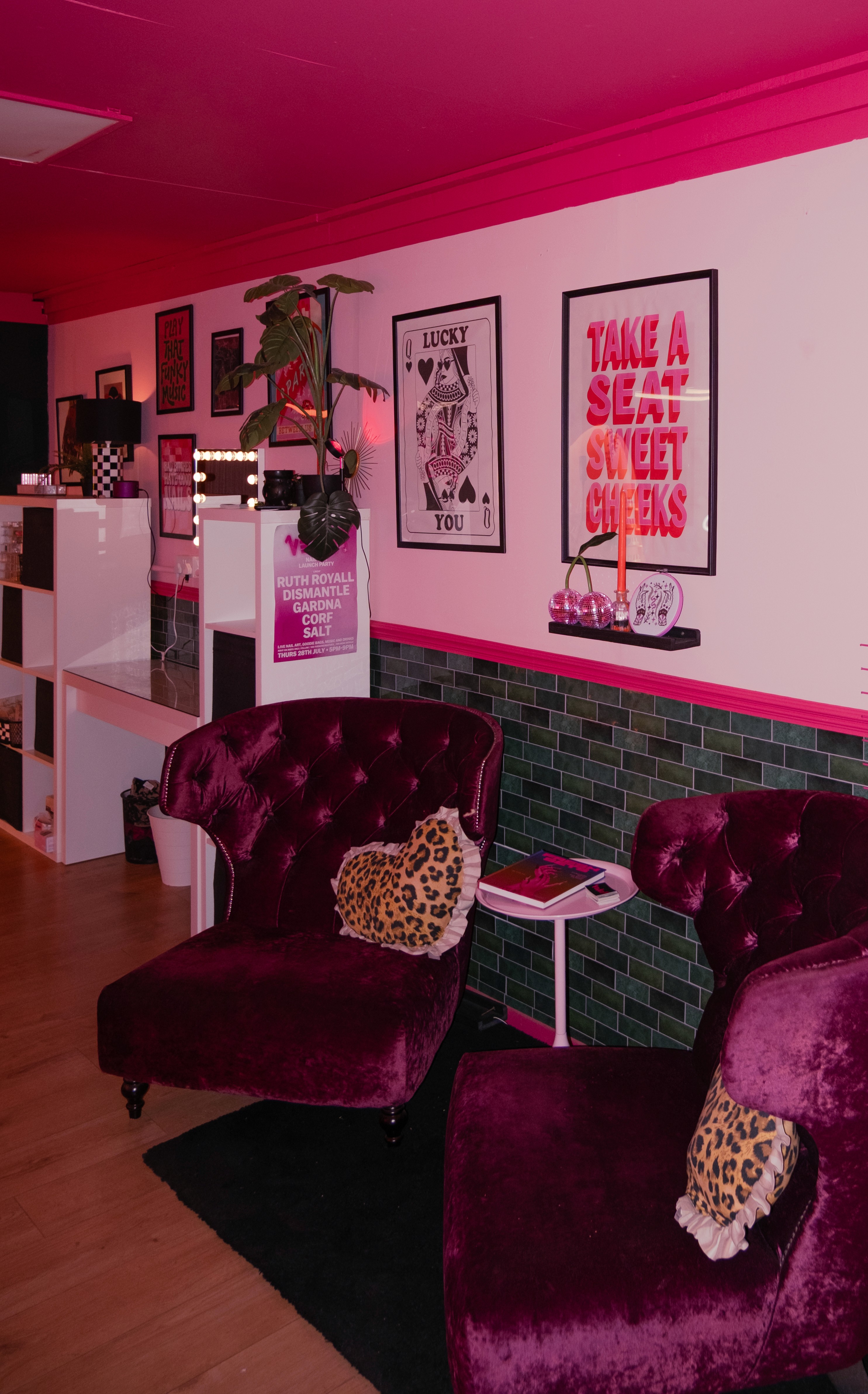 A wide shot of the salon’s interior featuring plush burgundy velvet wingback chairs, leopard-print heart pillows, and a gallery wall with posters like "Take a Seat Sweet Cheeks" and "Lucky You."