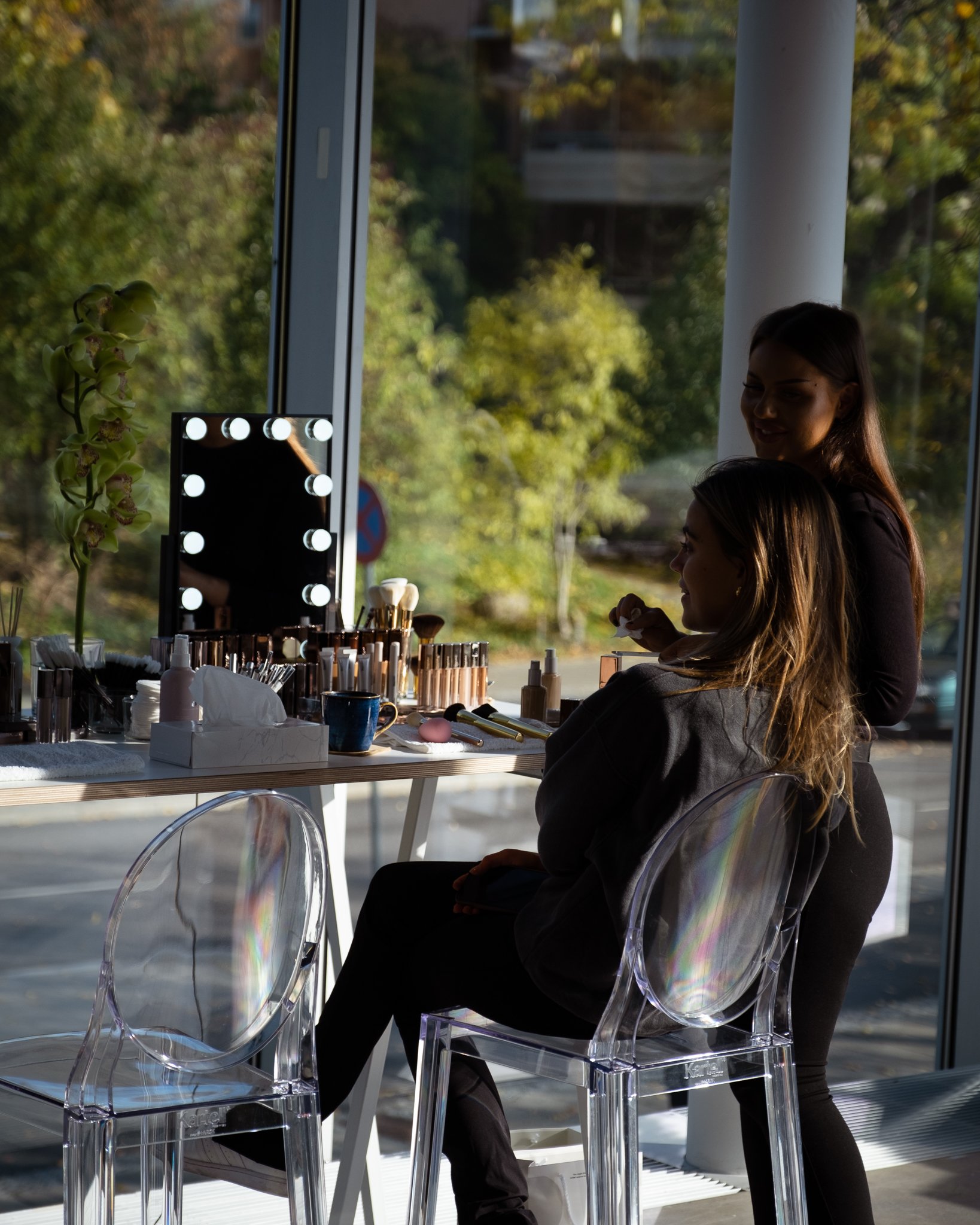 A makeup artist applying makeup to a woman seated at a vanity table with makeup products and brushes, with a large window showing a sunny outdoor scene of trees and greenery.