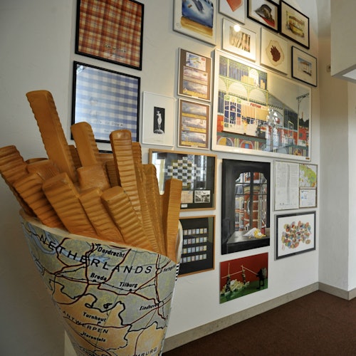 Egy nagy, élethű modell francia sültkrumpliból, melyet egy tölcsérben, a Hollandia térképével együtt helyeznek el, egy keretezett műalkotásokkal díszített fal mellett.