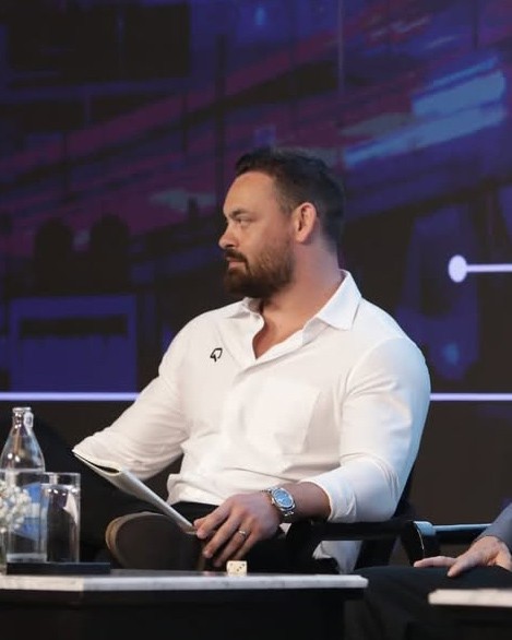 A stylish individual in a black cardigan and flat cap, paired with a white turtleneck, set against a neutral gray background.