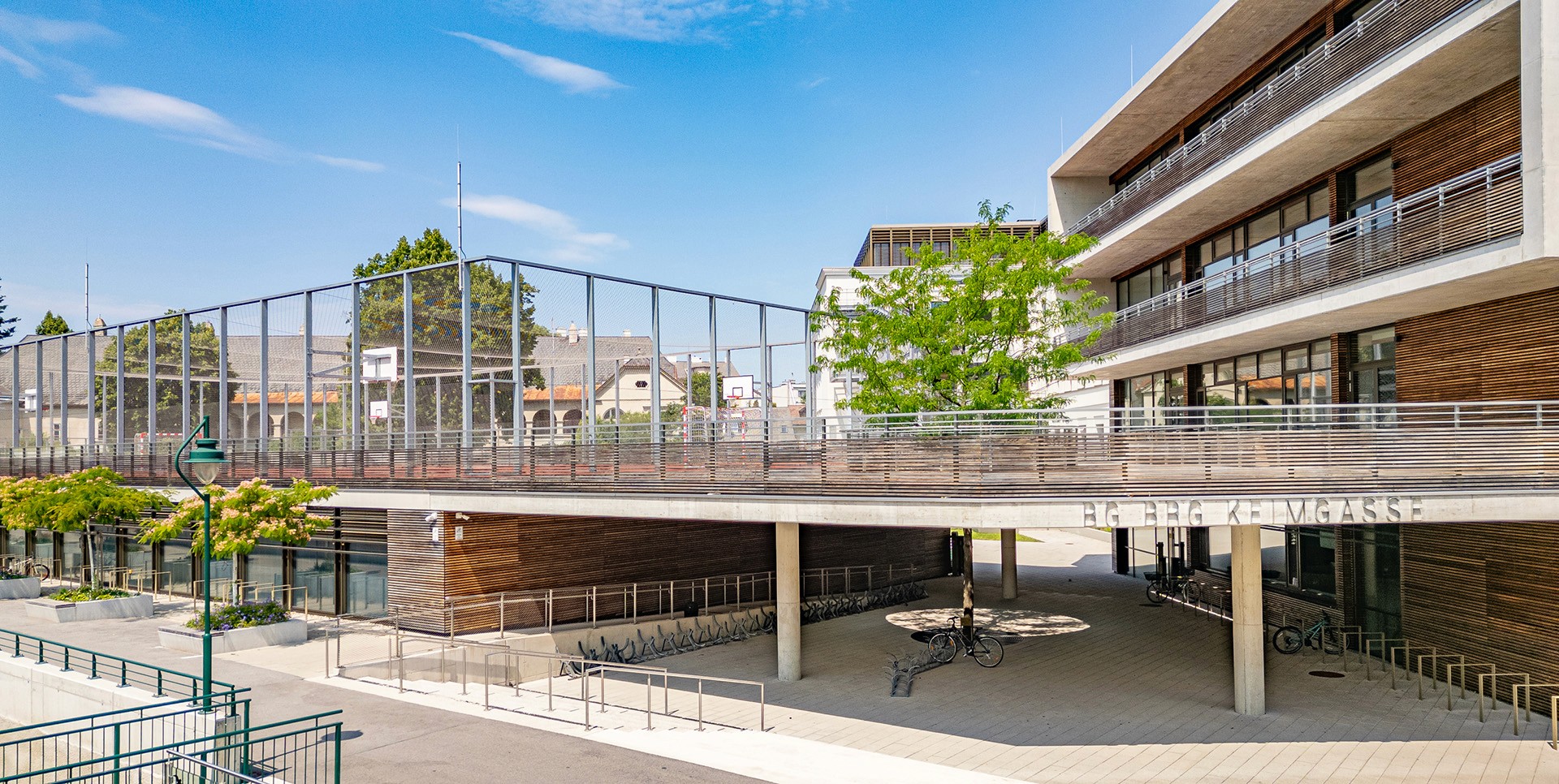 Moderner Schulhof mit Sportplatz, Gebäudekomplex und zentralem Baum als Gestaltungselement.