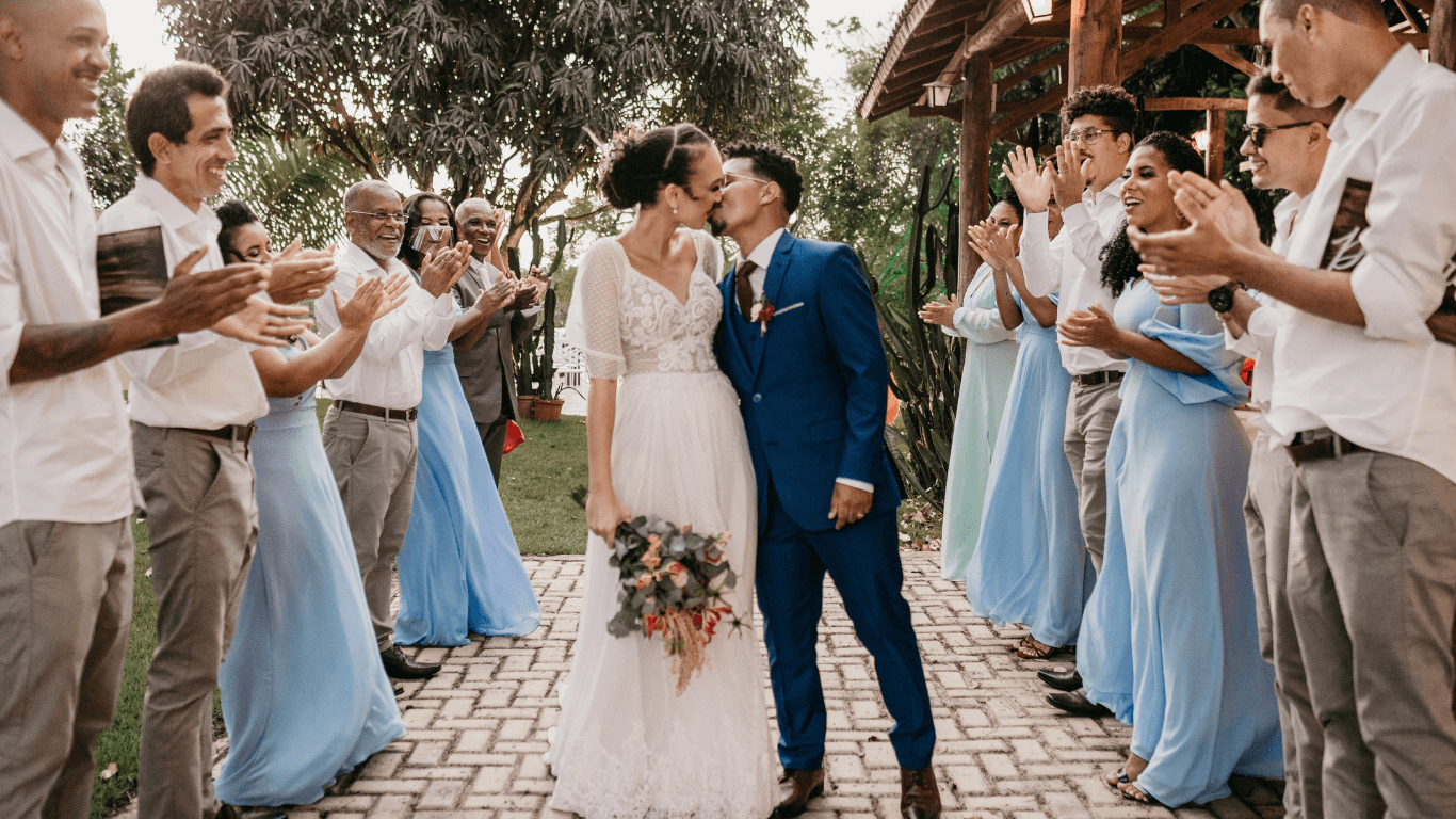 A newlywed couple shares a kiss surrounded by guests in formal attire, celebrating during their wedding.