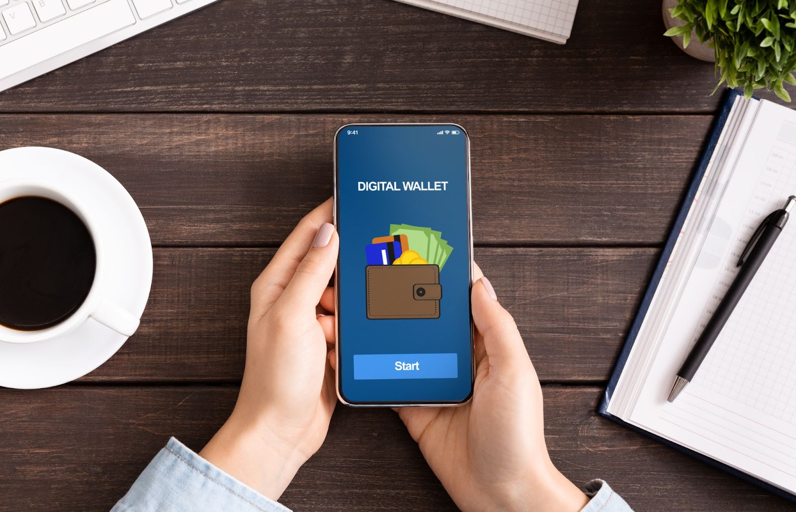 Woman holding a phone with the words “digital wallet” on the screen next to a cup of coffee, a notebook, a pen, and a plant. 