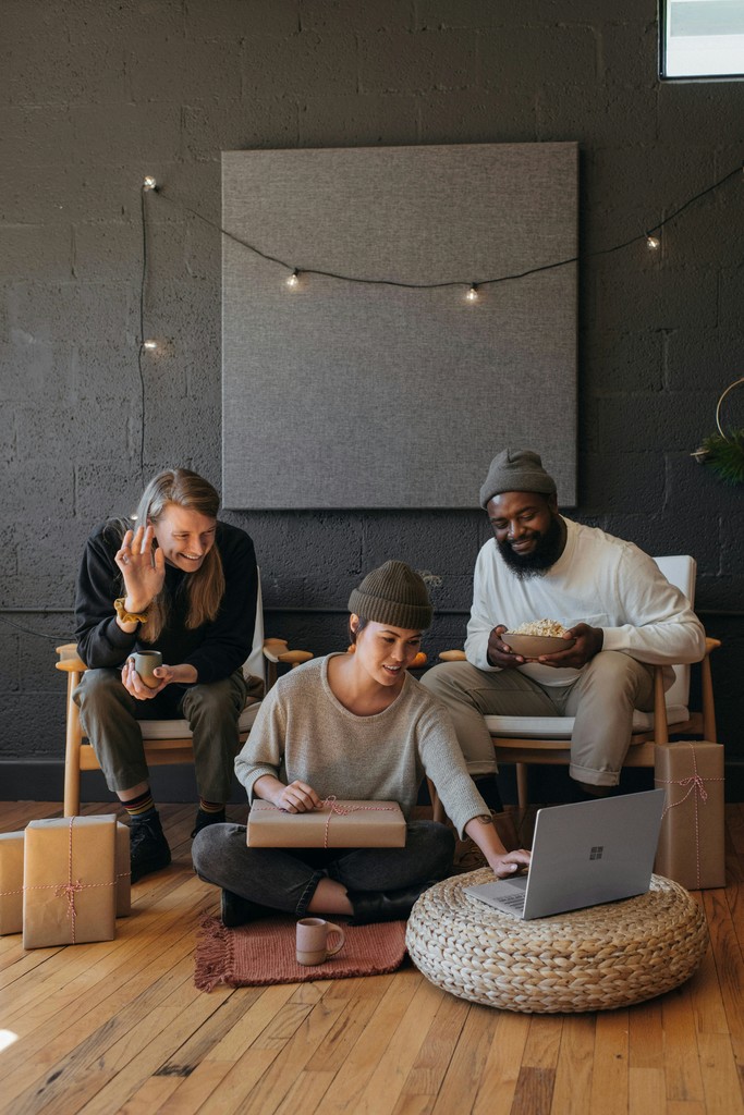 A group of friends enjoying a virtual gathering, with one waving at the laptop, another holding a wrapped gift, and another eating popcorn, creating a cozy and festive atmosphere.