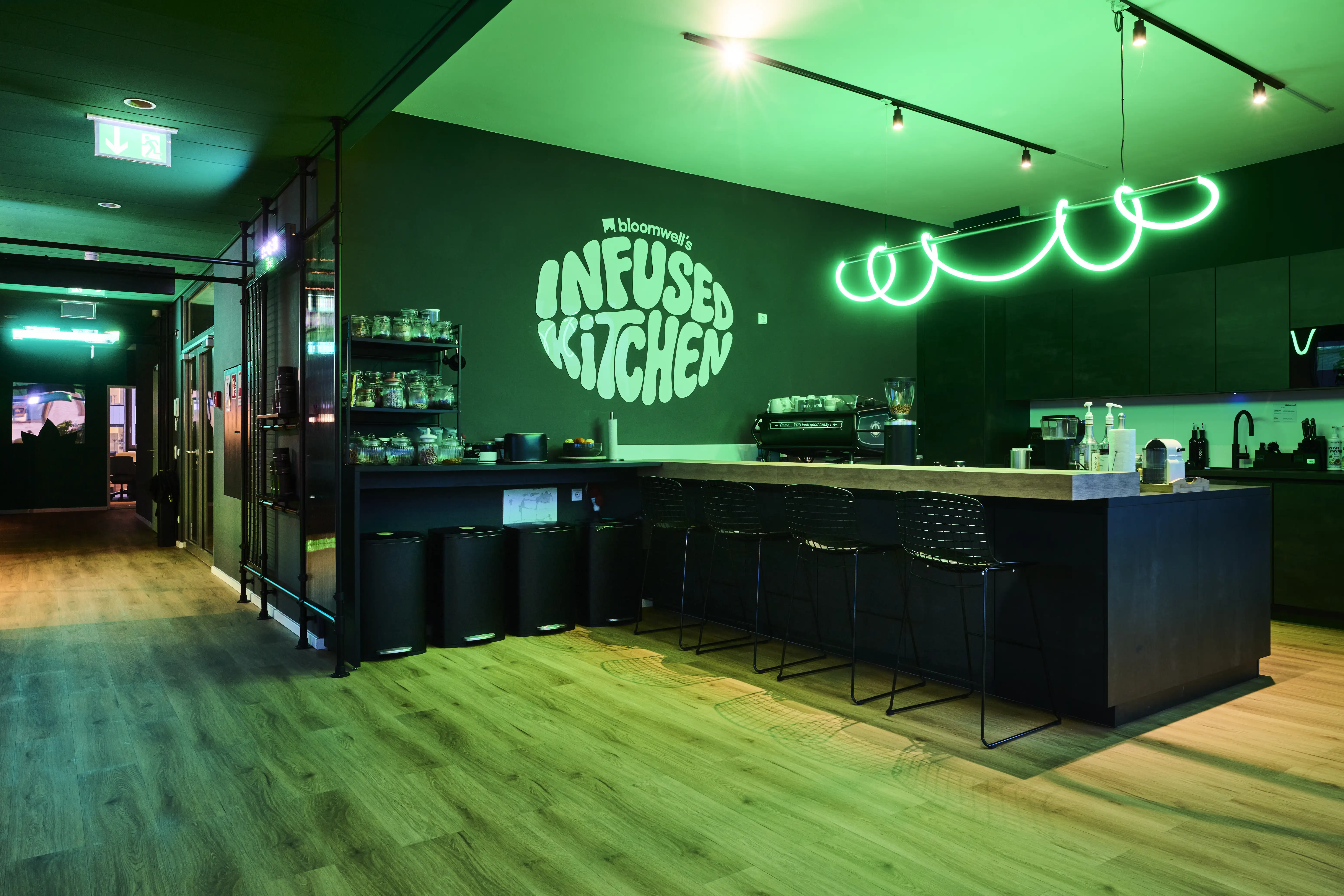 Dark kitchen at BLOOMWELL with black cabinets and bar stools, a green neon squiggle light, the Infused Kitchen logo glowing on a black wall and green-lit shelves.