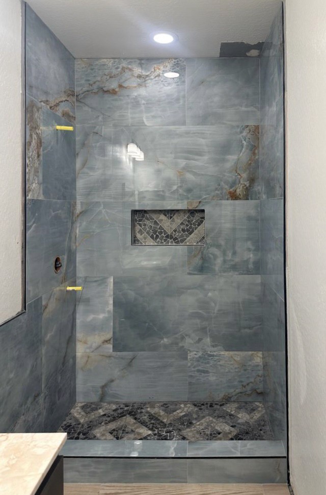 Shower wall tile installation in progress featuring large format blue-grey marble-look tiles and a built-in niche with a dark chevron mosaic tile pattern.