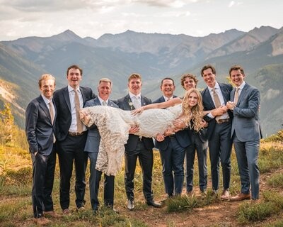 The groom and groomsmen laugh while holding the bride horizontally for a playful group portrait.