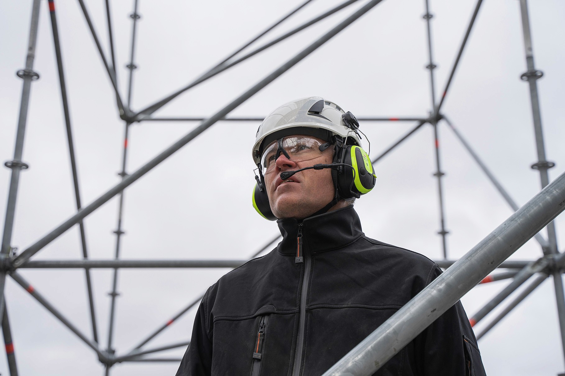 Construction worker wearing a helmet, safety glasses, and ear protection, looking up at scaffolding.