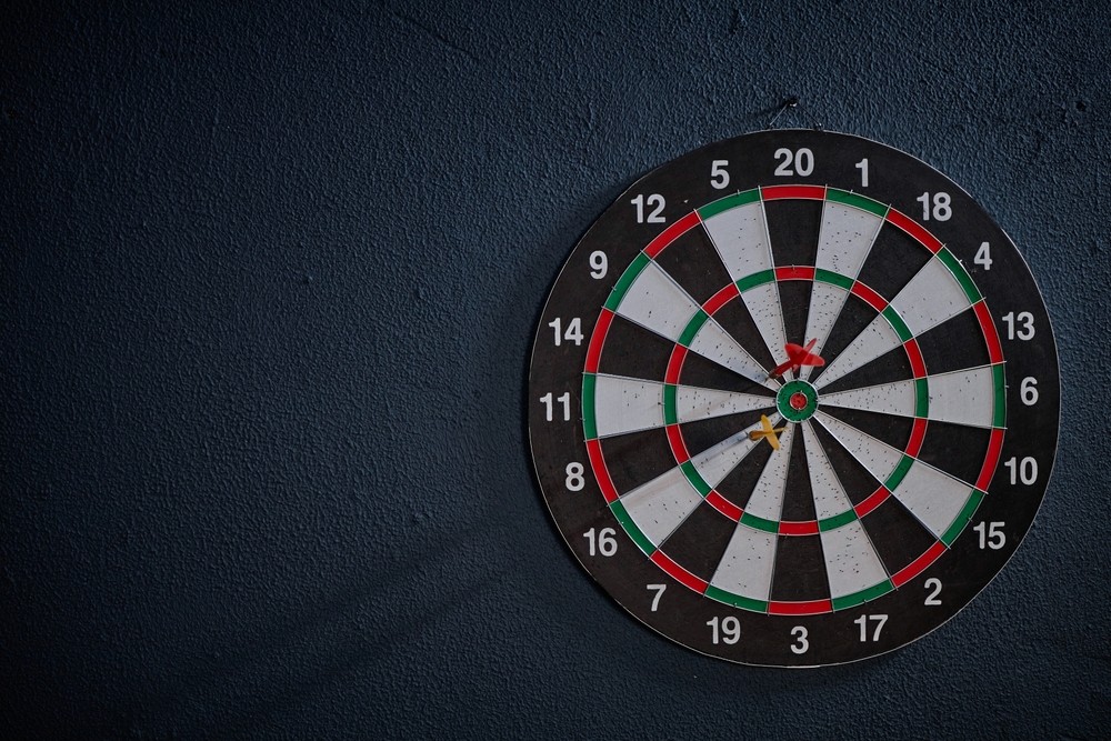 A young person with curly hair smiles at the camera while sitting in front of a laptop against a dark background.