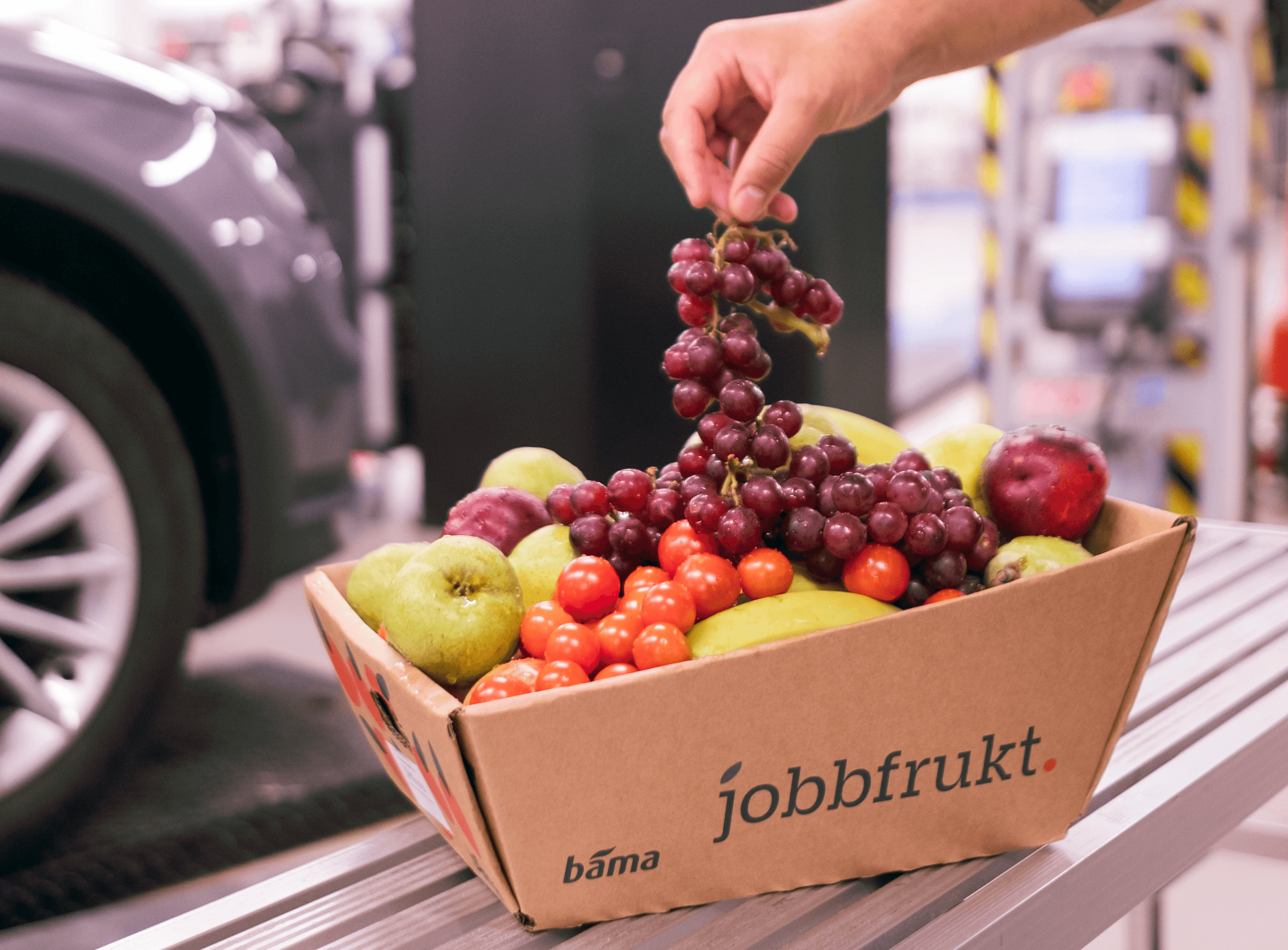 En hånd løfter en klase røde druer fra en Jobbfrukt-kurv fylt med epler, bananer, tomater og annen frisk frukt.