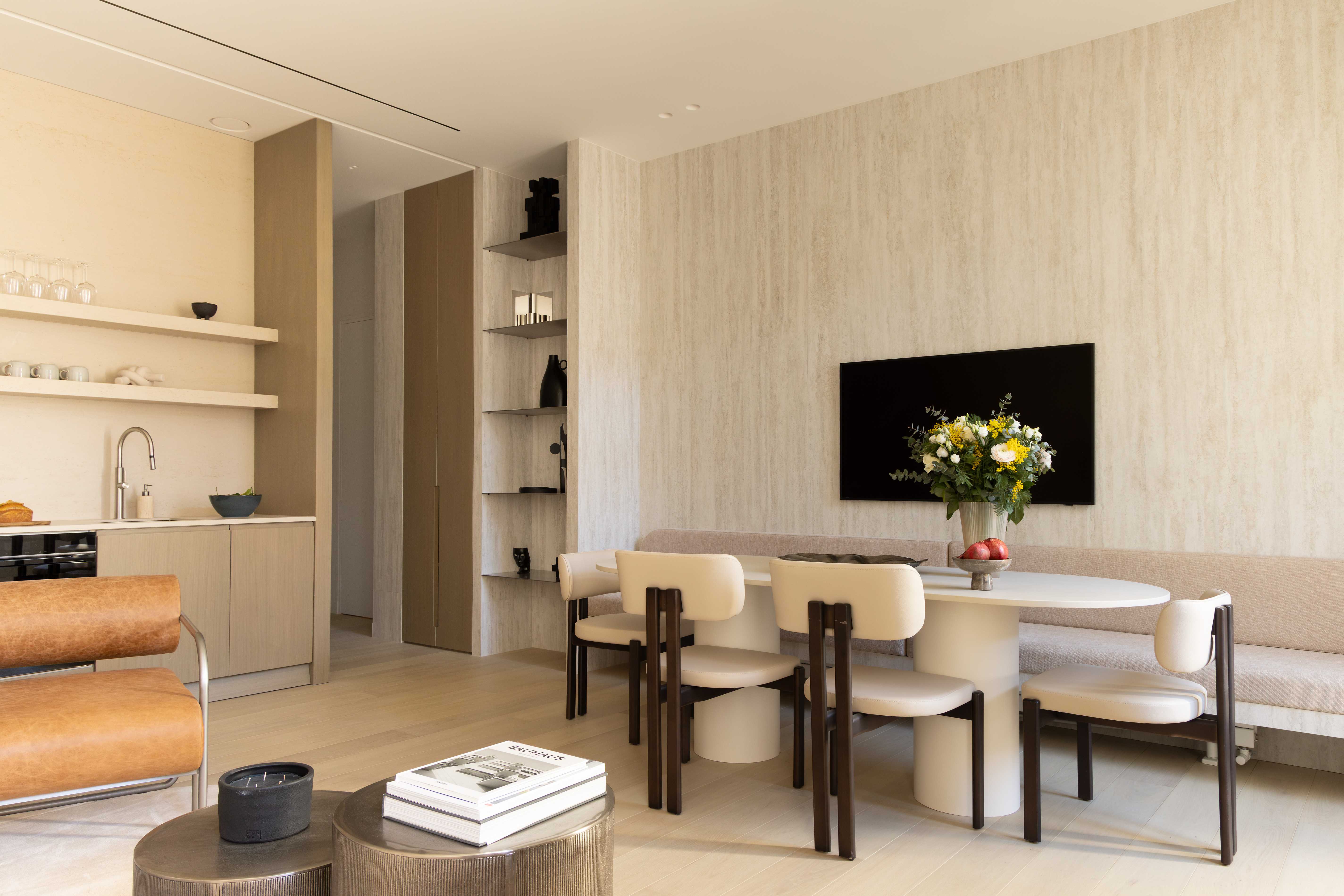 Vue d'ensemble salle a manger et cuisine, mur texture travertin, television, table ovale et etageres bois clair