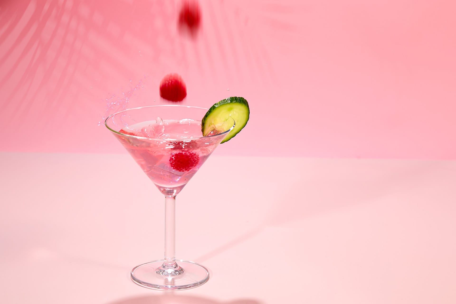 Orzeźwiający różowy koktajl ozdobiony plasterkiem ogórka i malinami wpadającymi do drinka, tworzącymi plusk na miękkim różowym tle.