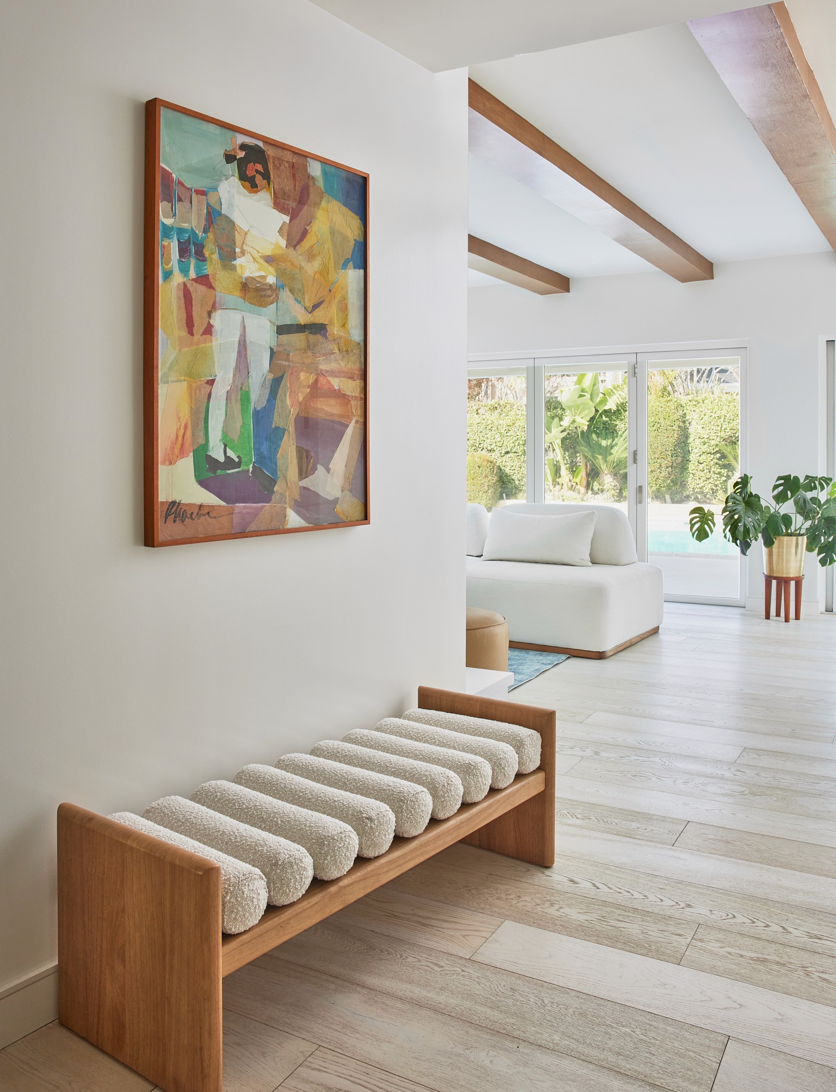 Bright front entryway with colorful art and white textured bench with a peek into the family room and backyard through bi-fold doors by Jubilee Interiors in Los Angeles, California