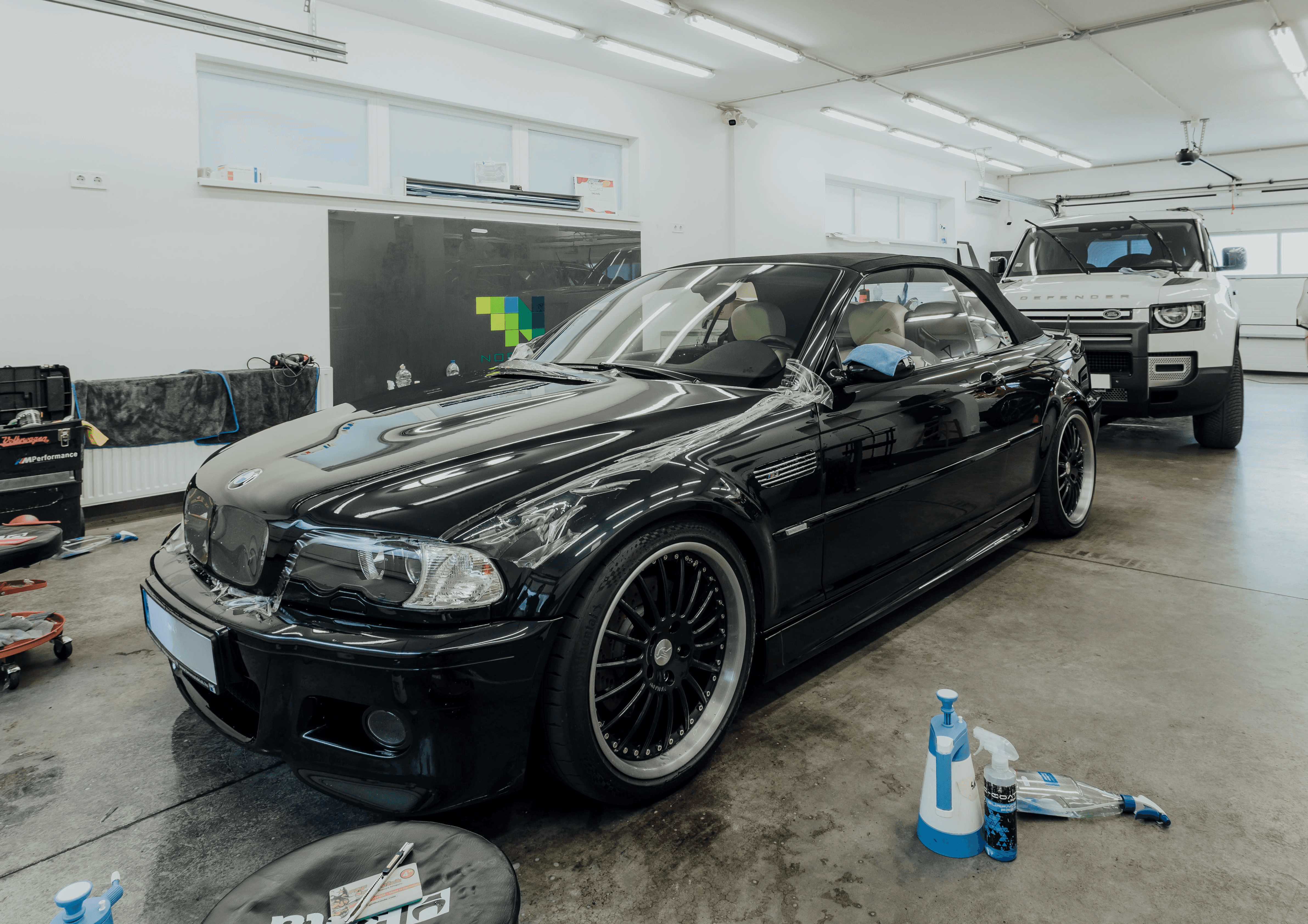 Teljes felújításon átesett, mélyfekete BMW E46 M3 Cabriolet a Nortek budapesti műhelyében. Az ikonikus youngtimer sportautó épp front kővédő fóliát (PPF) és ClearPlex szélvédővédő fóliát kap a gyári állapot megőrzése érdekében. A háttérben egy fehér Land Rover Defender látható.