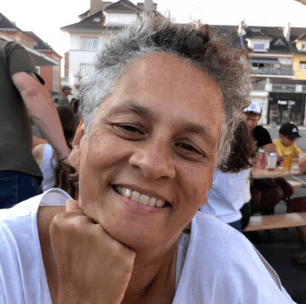 Smiling female therapist with short gray hair, wearing a brown top and a purple shawl, surrounded by a cozy background.
