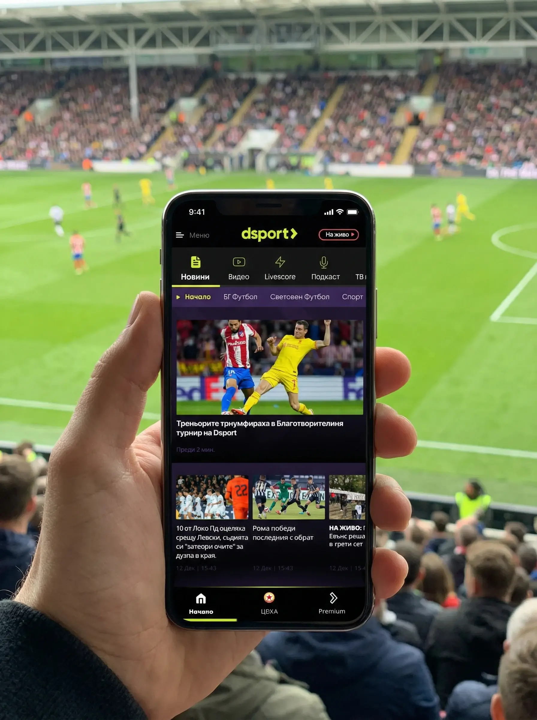 A person holds a smartphone displaying a sports news app with a live soccer match in the background at a stadium.