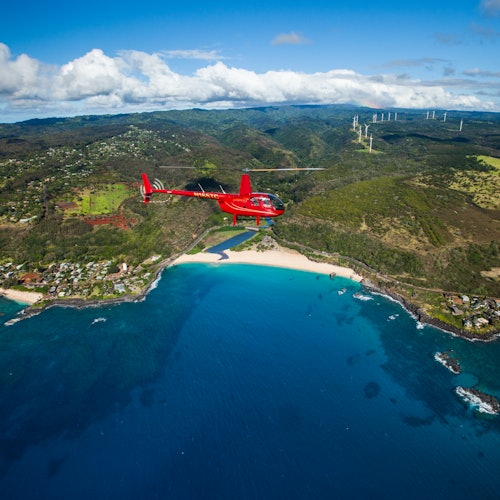 เฮลิคอปเตอร์สีแดงบินอยู่เหนือภูมิทัศน์ชายฝั่งที่มีน้ำสีฟ้าคราม ชายหาดที่มีทราย บ้านเรือน เนินเขา และกังหันลมในระยะไกล