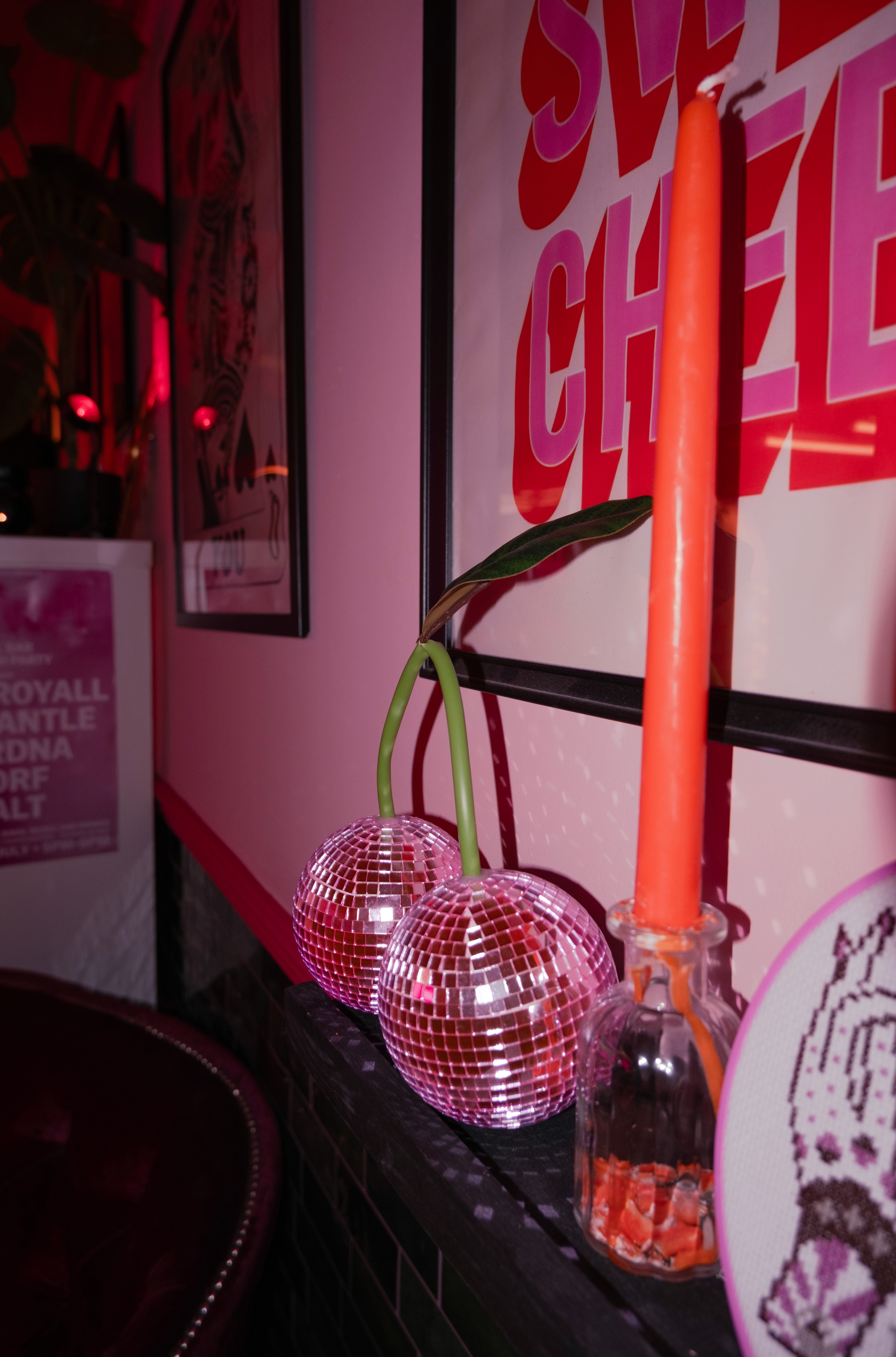 A whimsical decor detail featuring two pink disco-ball "cherries" with green stems sitting on a shelf next to an orange taper candle in a glass holder.
