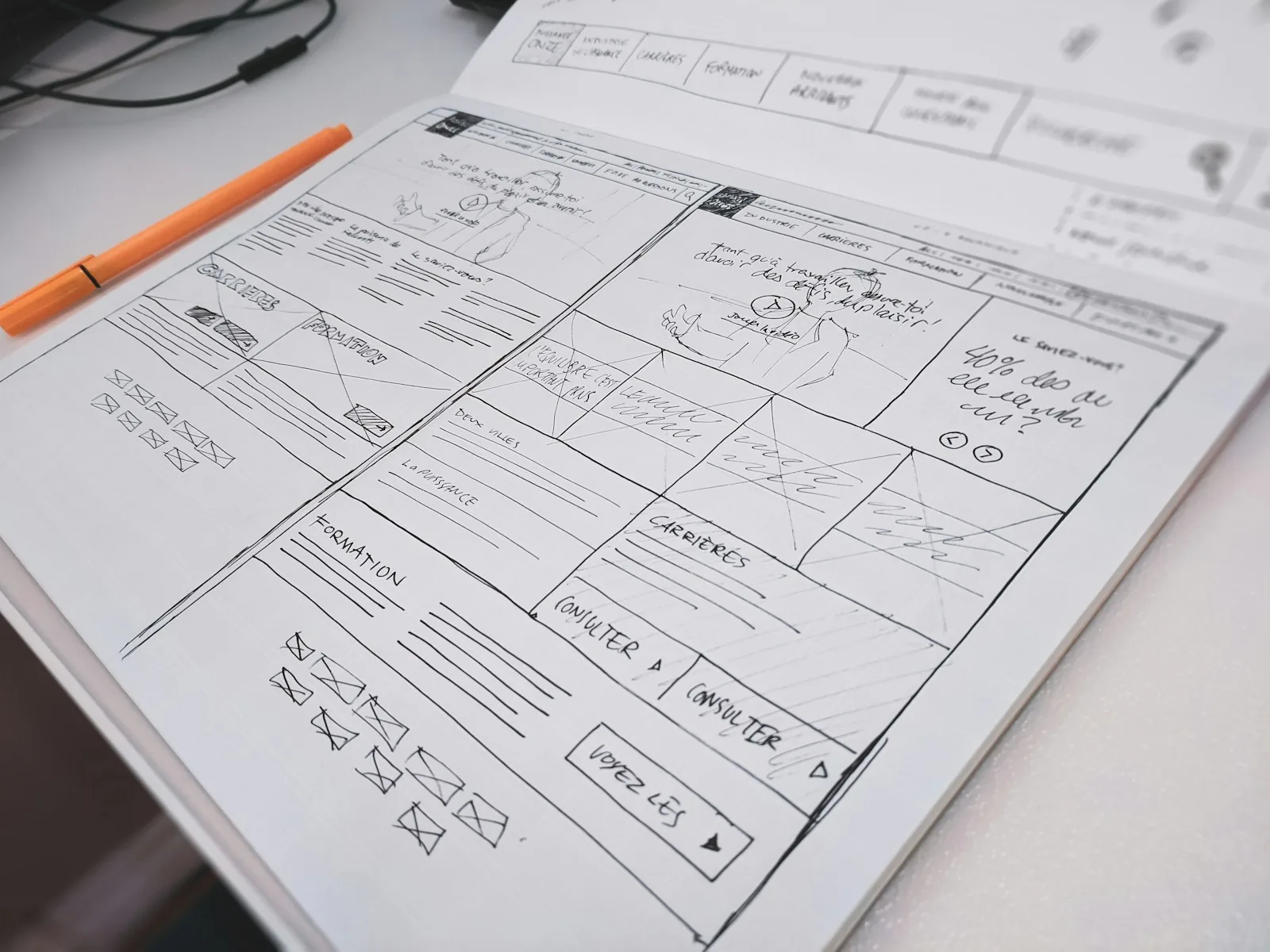 Hand-drawn website wireframes in an open sketchbook, with boxes, crossed image placeholders and button labels, next to an orange pen