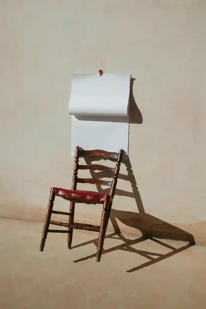 A wooden chair with a distressed red seat stands against a beige wall. Above the chair, a minimalist design hangs, casting a shadow that adds depth to the composition and creating a modern aesthetic.