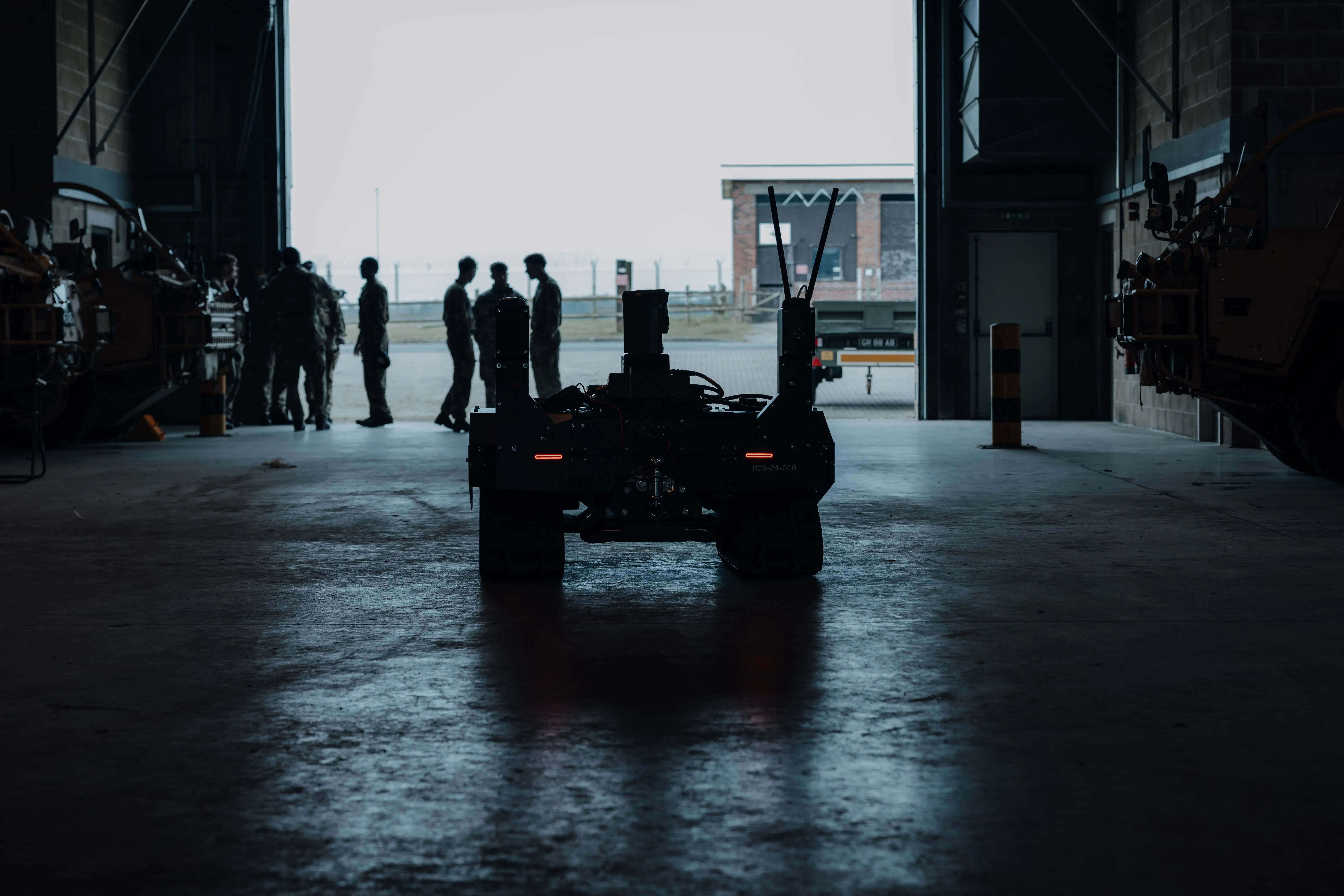 A silhouette of military personnel and a vehicle inside a large, dimly lit hangar, with light streaming in from an open entrance, highlighting the figures and creating dramatic shadows.