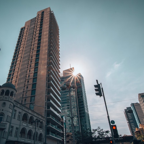 Tall buildings in a cityscape with the sun peeking between them, traffic lights, and power lines against a clear sky.