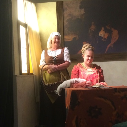 Two women in period clothing inside a dimly lit room, one seated at a table writing with a quill, and the other standing, smiling.