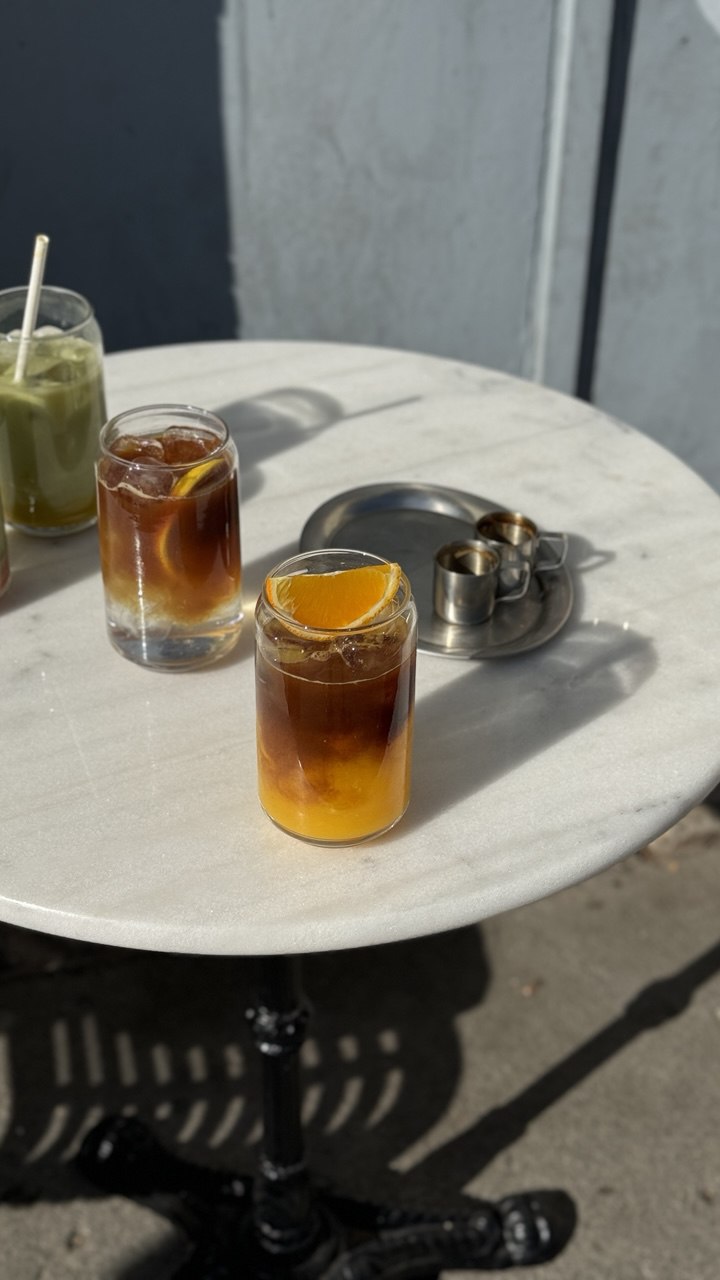 Two cold brew coffee drinks with ice and orange slices are served in clear glasses on a round white marble table, accompanied by a stack of napkins and a partial view of another green beverage.