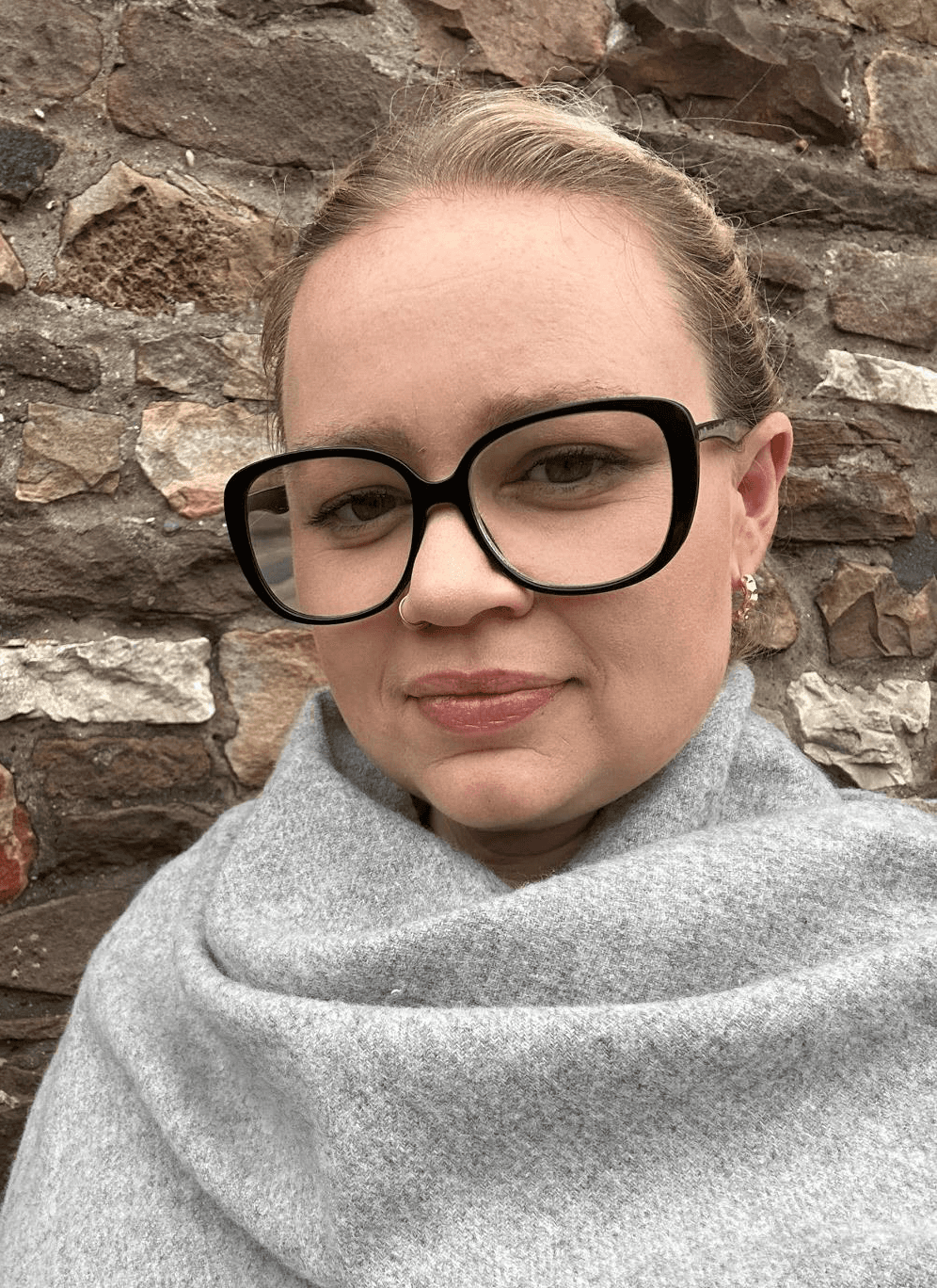A woman wearing a grey shawl and black rimmed glasses standing against a stone wall. 