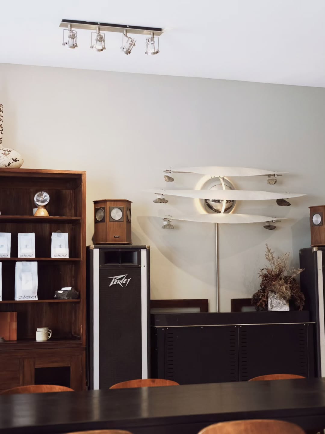 A modern interior featuring a wooden shelf with coffee bags, vintage clocks, and decorative elements, along with tall speakers and a unique wall-mounted sculpture resembling airplane propellers.