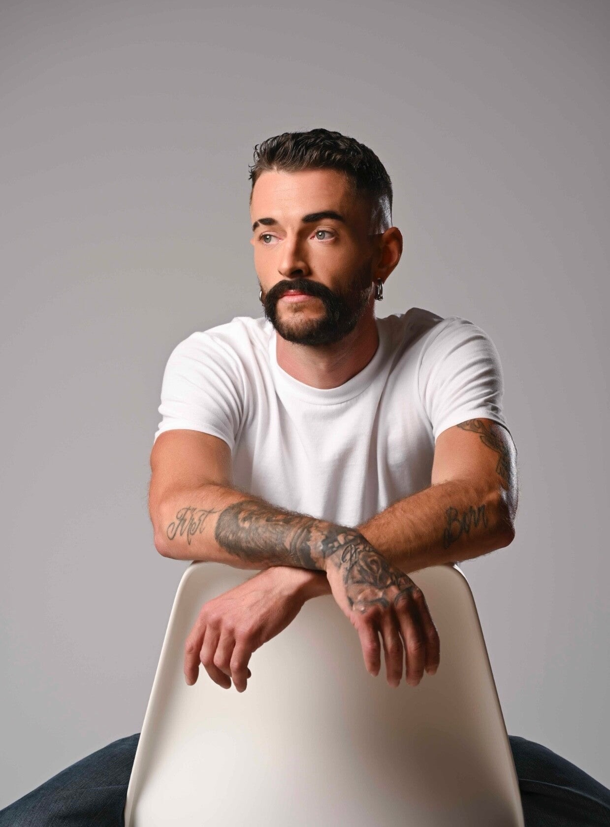 Tattooed bearded man in white T-shirt sitting backward on chair against neutral studio background.