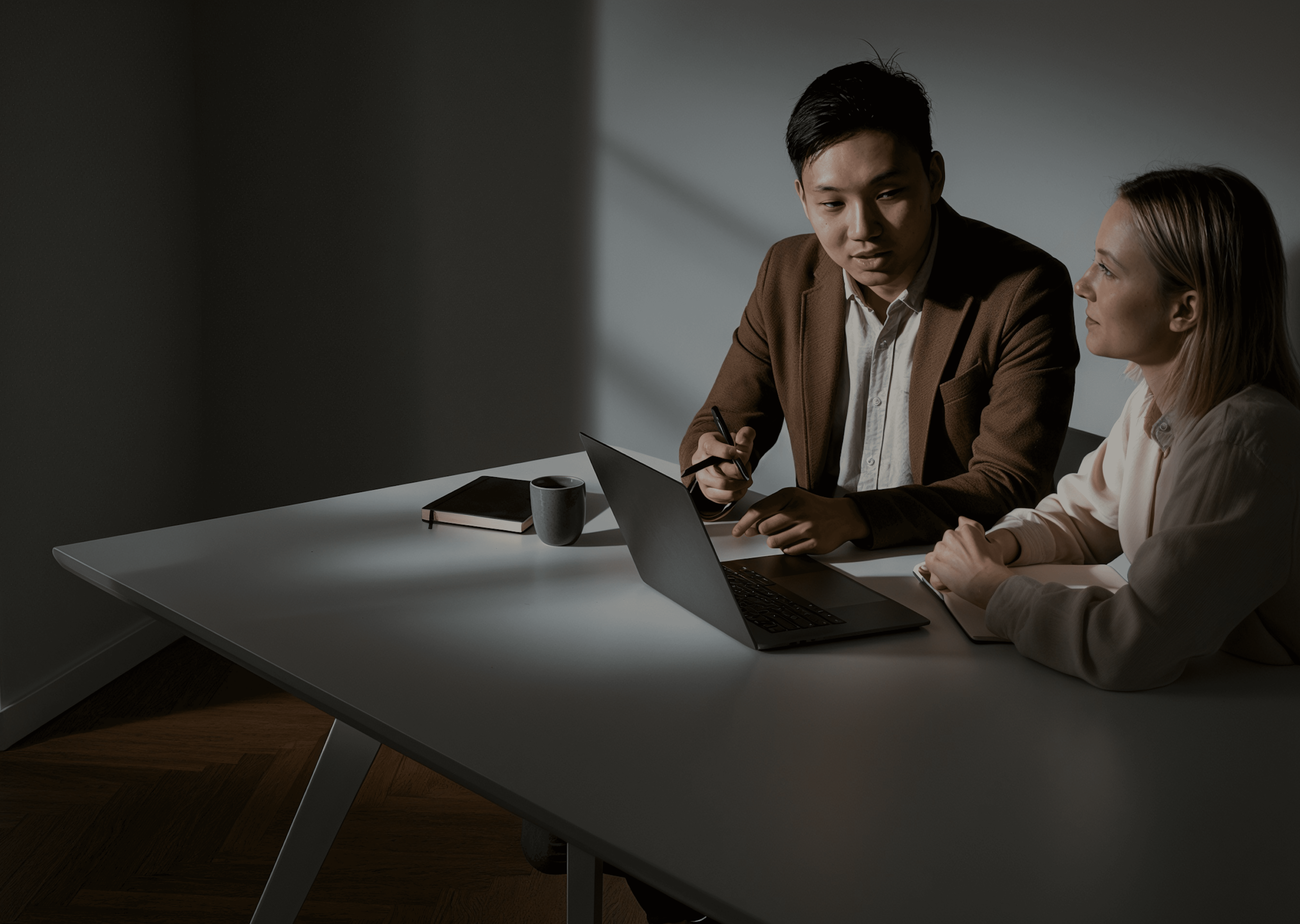 Two colleagues talking at a laptop in a dim office.