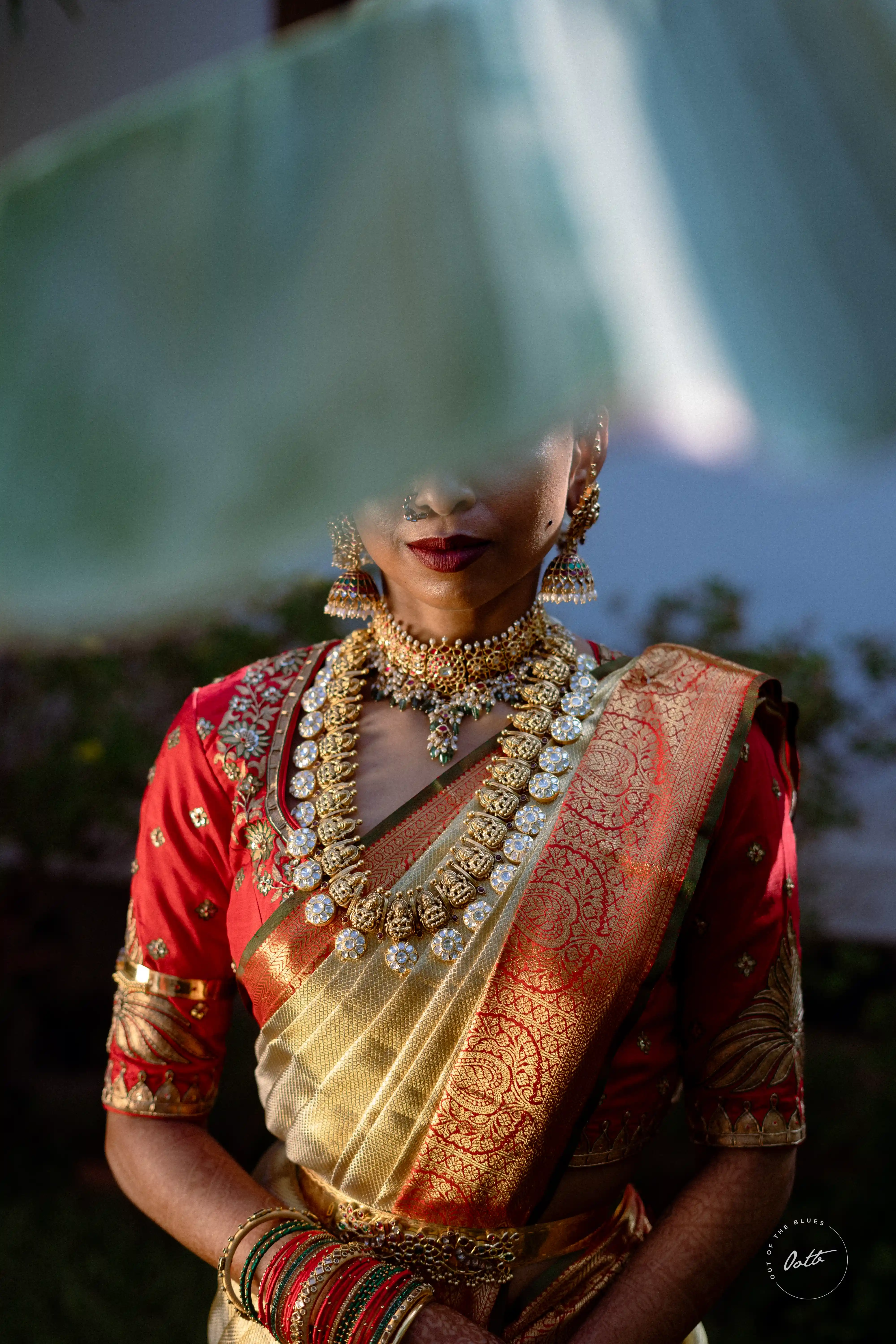 Fine art shot of a bride dressed in intricate gold jewelry and traditional attire, her face partially obscured by leaves, creating a dreamlike effect by Out of The Blues Fine Art Wedding Photography in Hyderabad.