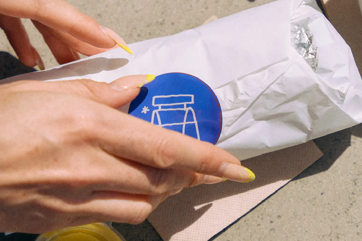Burrito wrapped in paper with blue Benchside Burritos sticker, a woman removing the sticker.