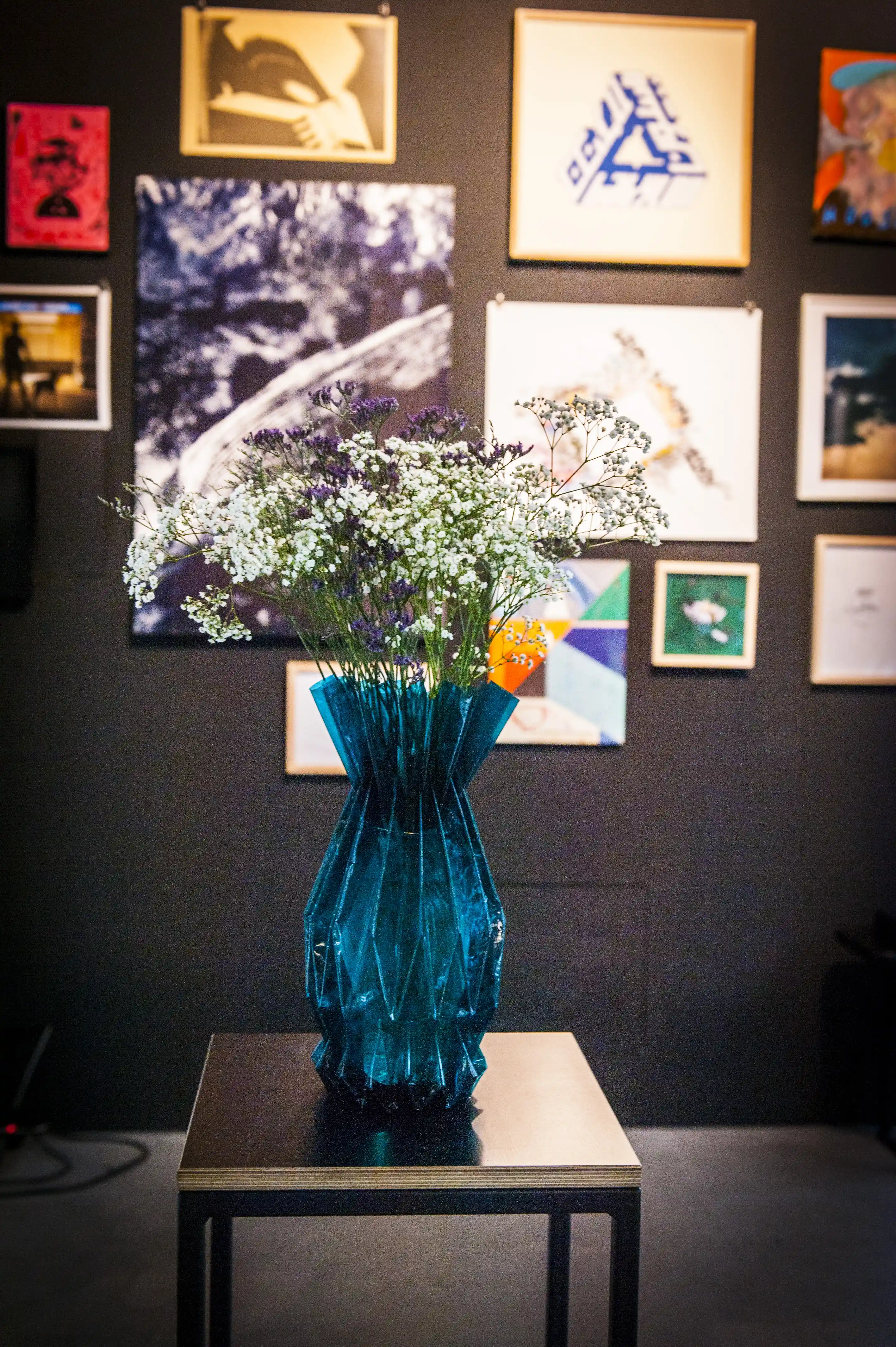 Faceted blue glass vase with wildflowers on a black side table in front of the salon-style artwork wall at the STUDIOLO gallery.