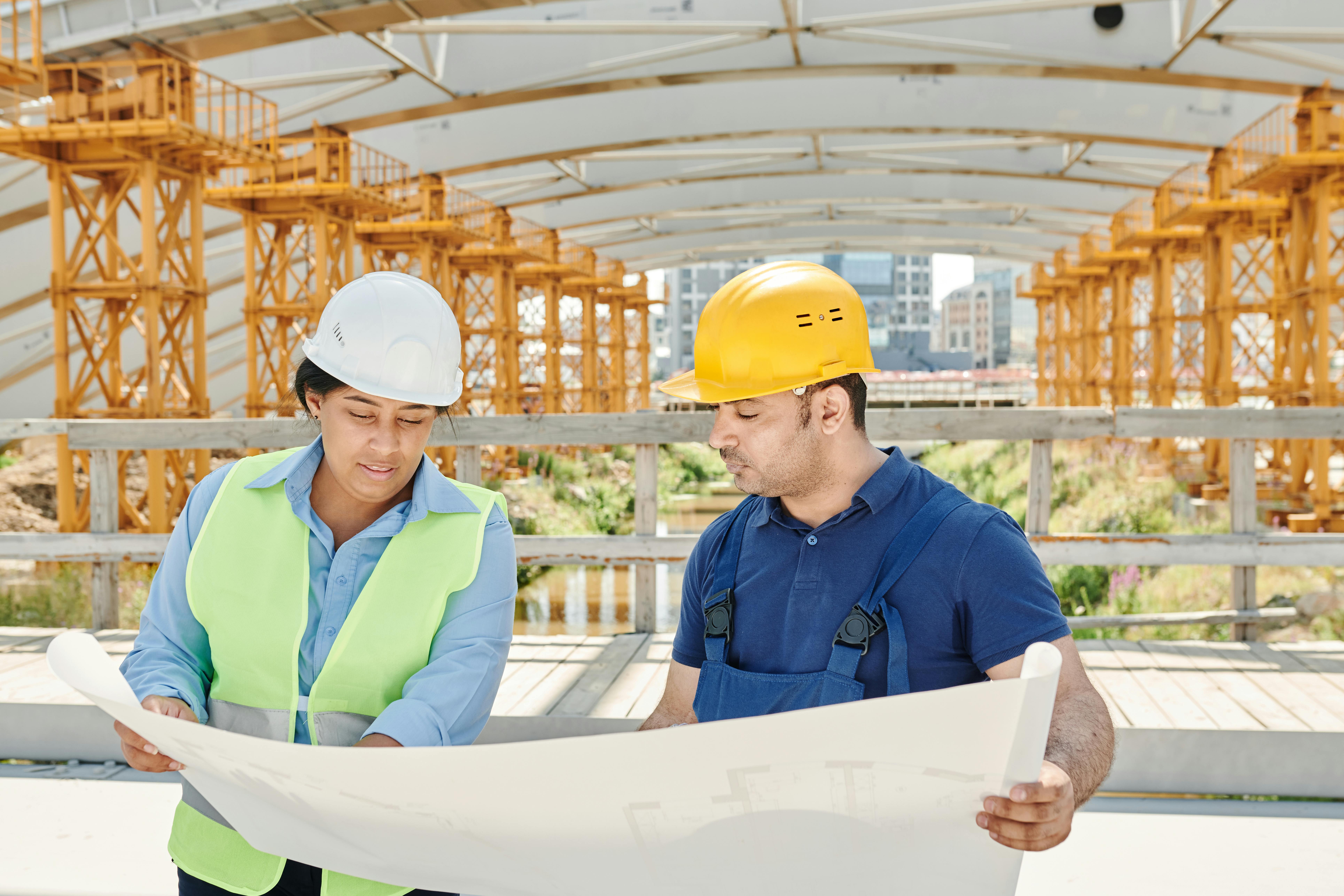 Two construction workers wearing hard hats review blueprints at a construction site. Steel beams in the background indicate an industrial setting.
